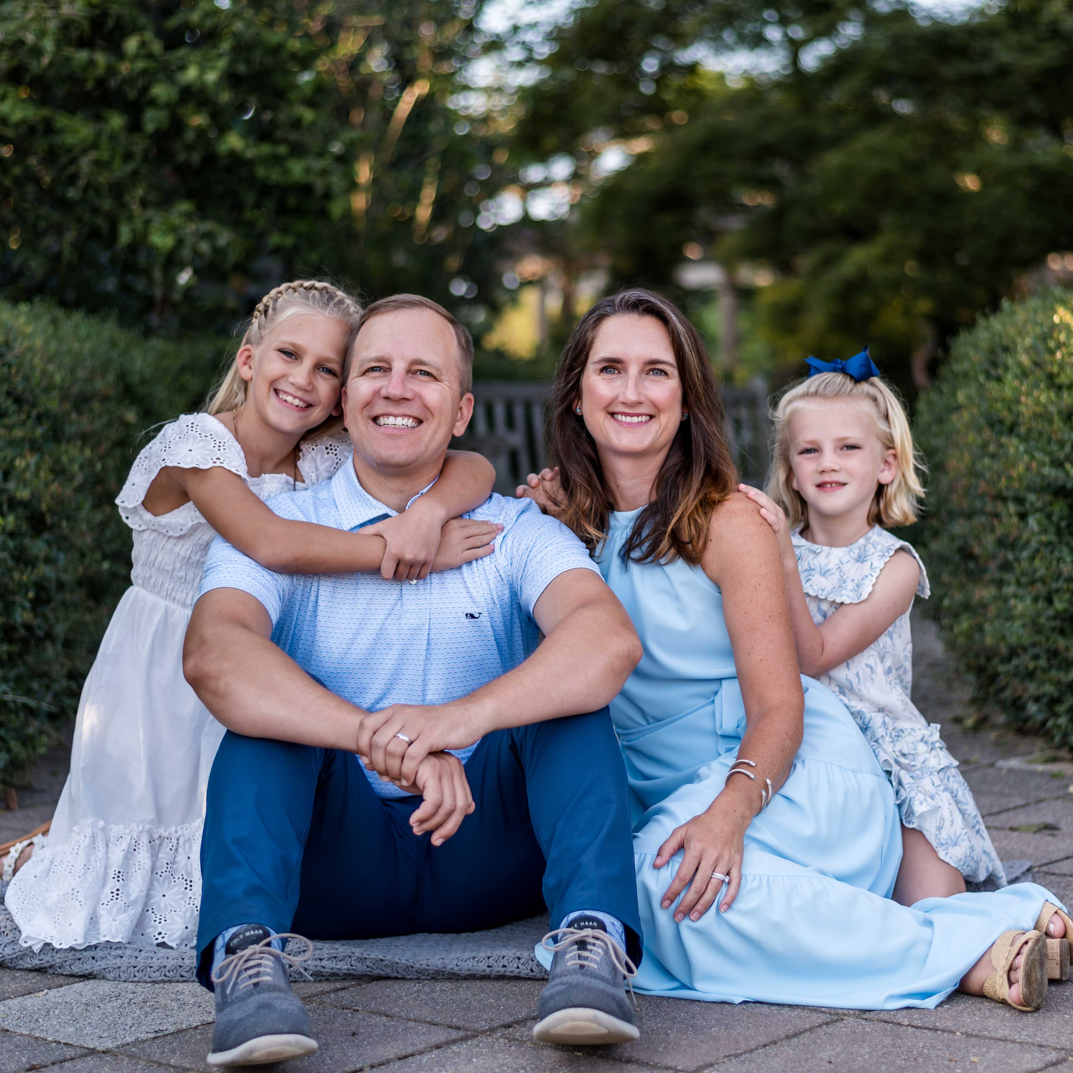 Nick and his family smile. His wife sits to the right and his daughters flank both sides. They all wear shades of white and blue.