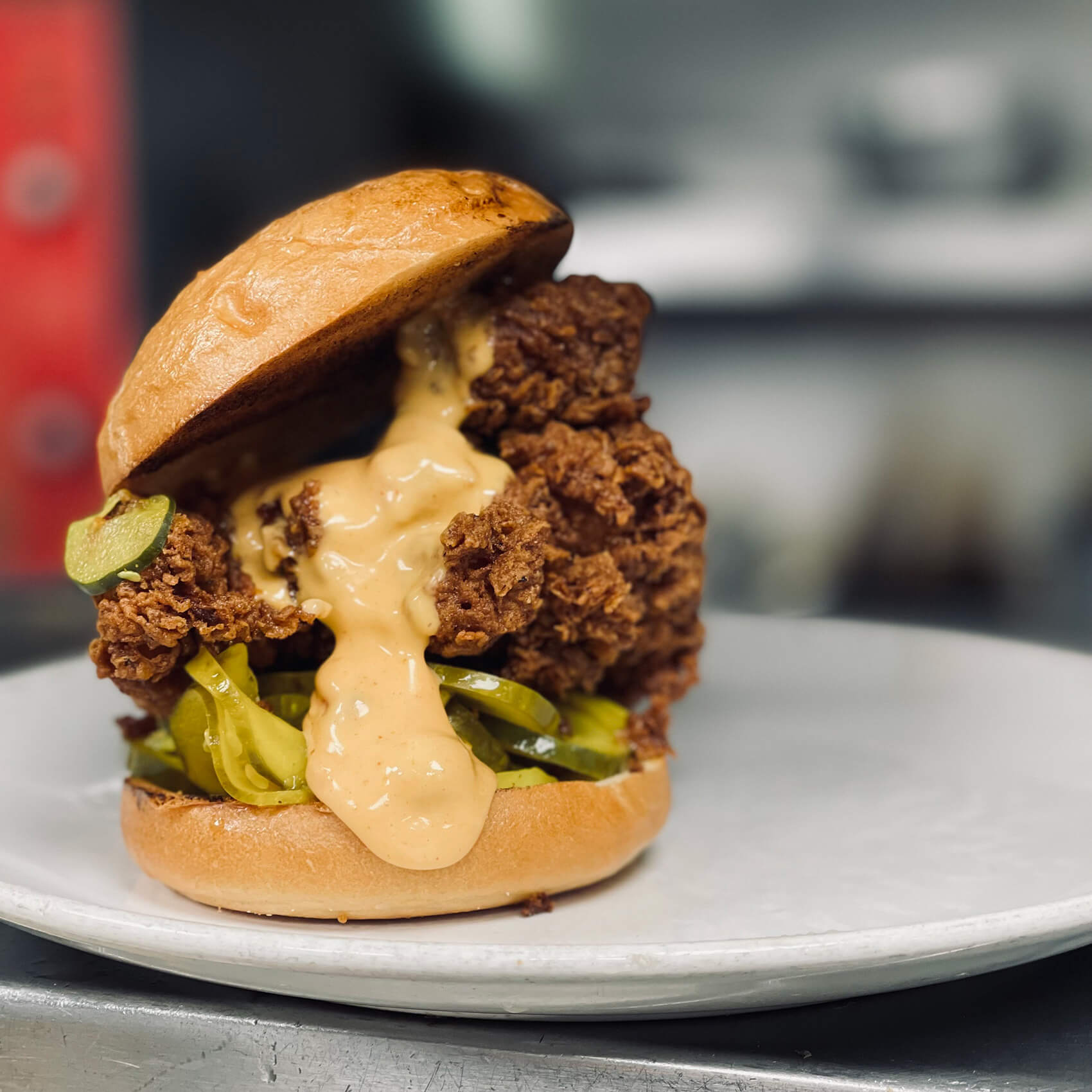 Close-up of The Bayou’s Nashville Fried Chicken Sandwich featuring a golden-fried chicken thigh topped with creamy pimento ranch sauce and pickles on a toasted bun, plated in a restaurant kitchen.