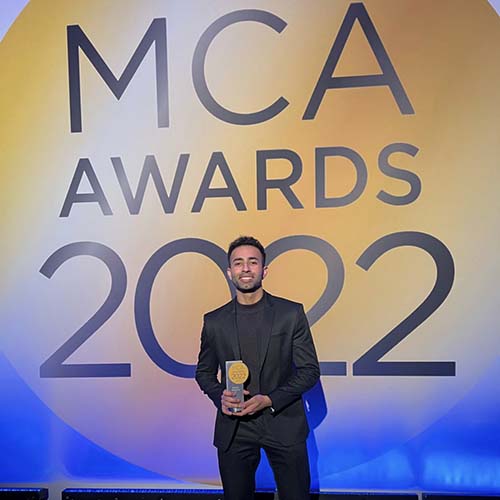Aakash Phulwani in a black sport coat and shirt holds an award at the MCA Awards 2022