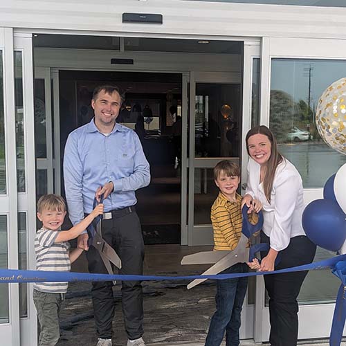 John and his wife hold big scissors to cut a ribbon with their sons at a newly opened hotel