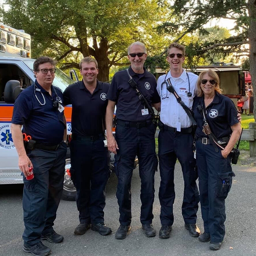 Five EMTs stand with stethoscopes around their necks and walkie talkies on their belts outdoors beside an ambulance on a warm day.