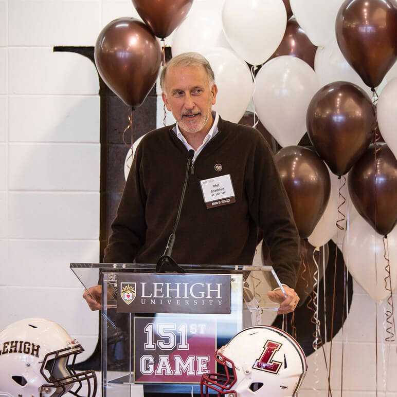 Man wearing a Lehigh name tag on his brown quarter-zip fleece stands at a podium with brown and white balloons behind him.