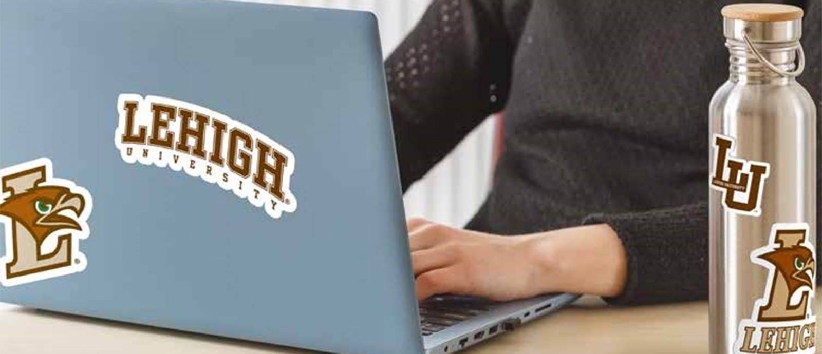 A close up of a person at a desk with a laptop and silver travel bottle adorned with Lehigh University stickers.