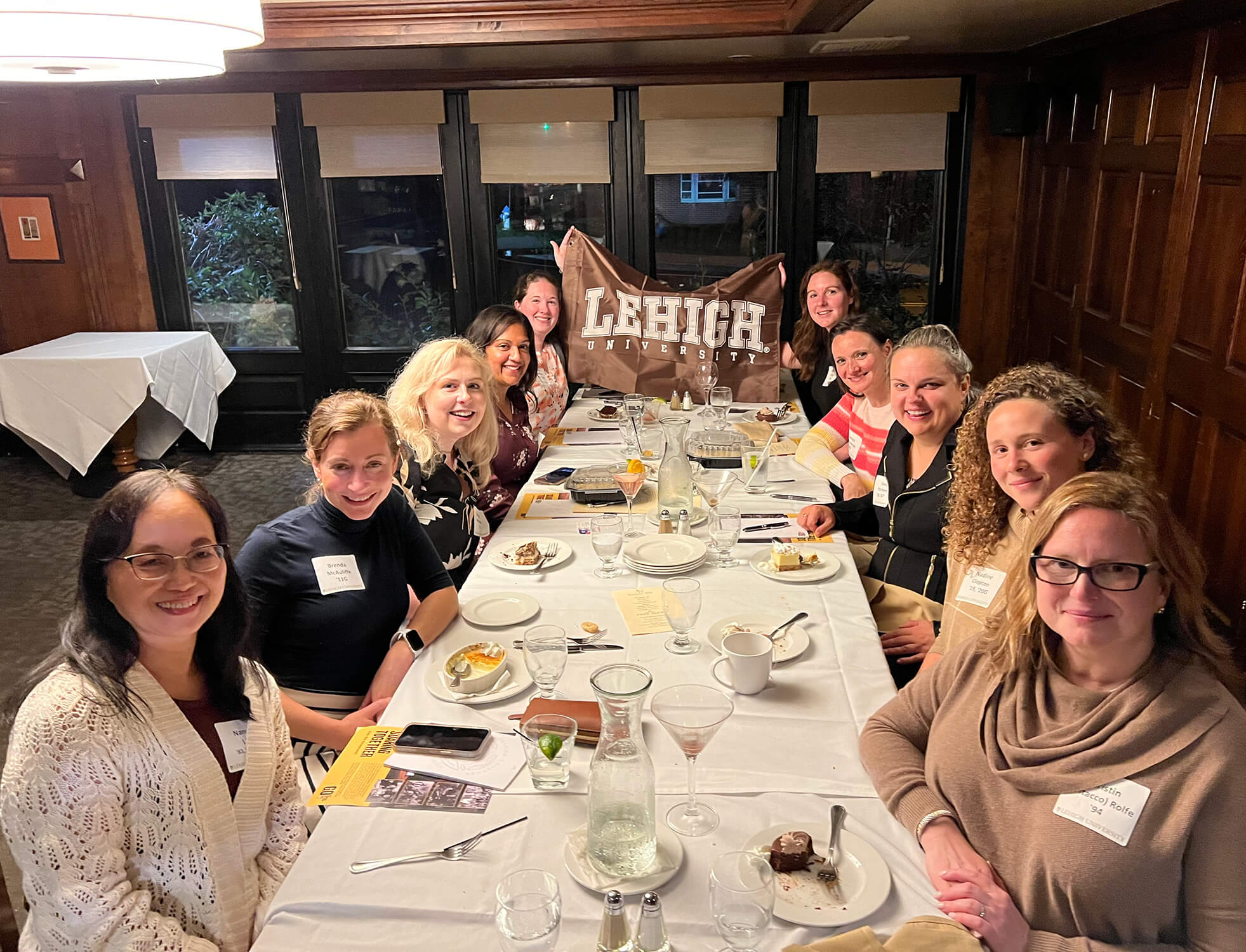 A long table of women pose for a photo with two at the far end holding up a Lehigh flag.