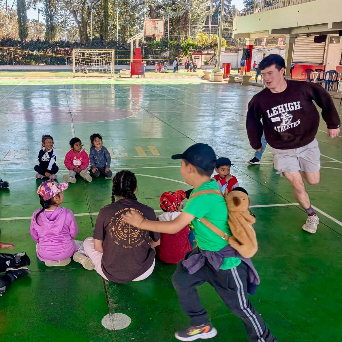 Wearing a brown Lehigh Athletics sweatshirt, Nate Raney chases a child around a circle as they play in the shade.