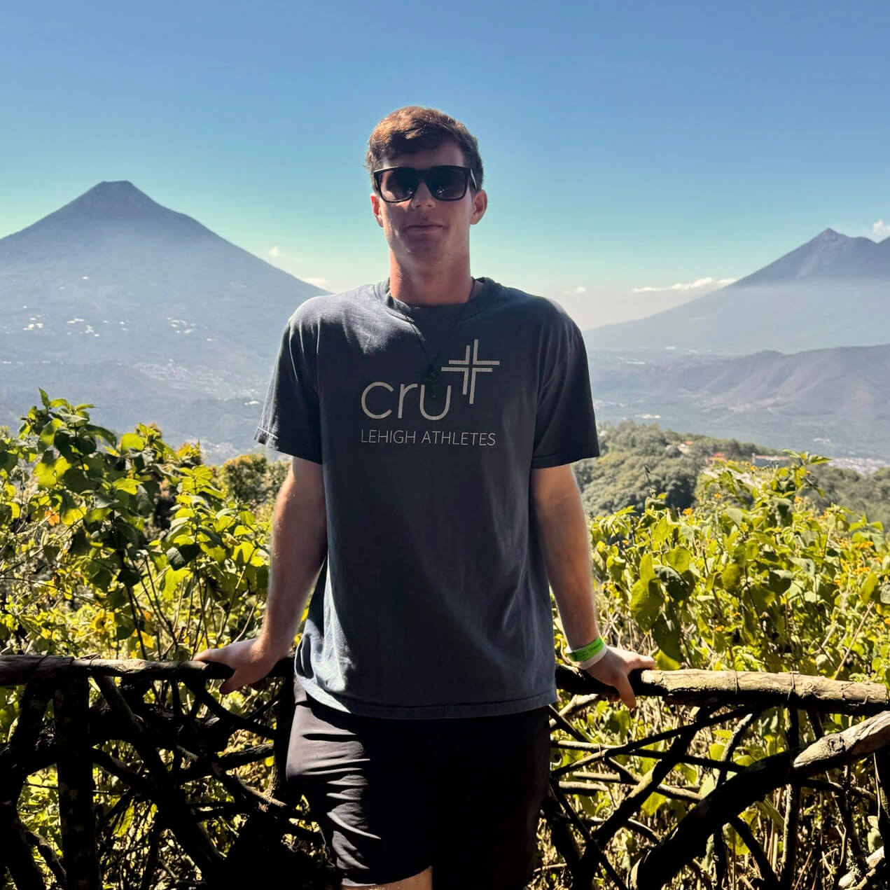 Nate Raney stands against a fence overlooking the green, mountainous landscape of Guatemala, wearing a shirt that reads “Lehigh Athletes.”