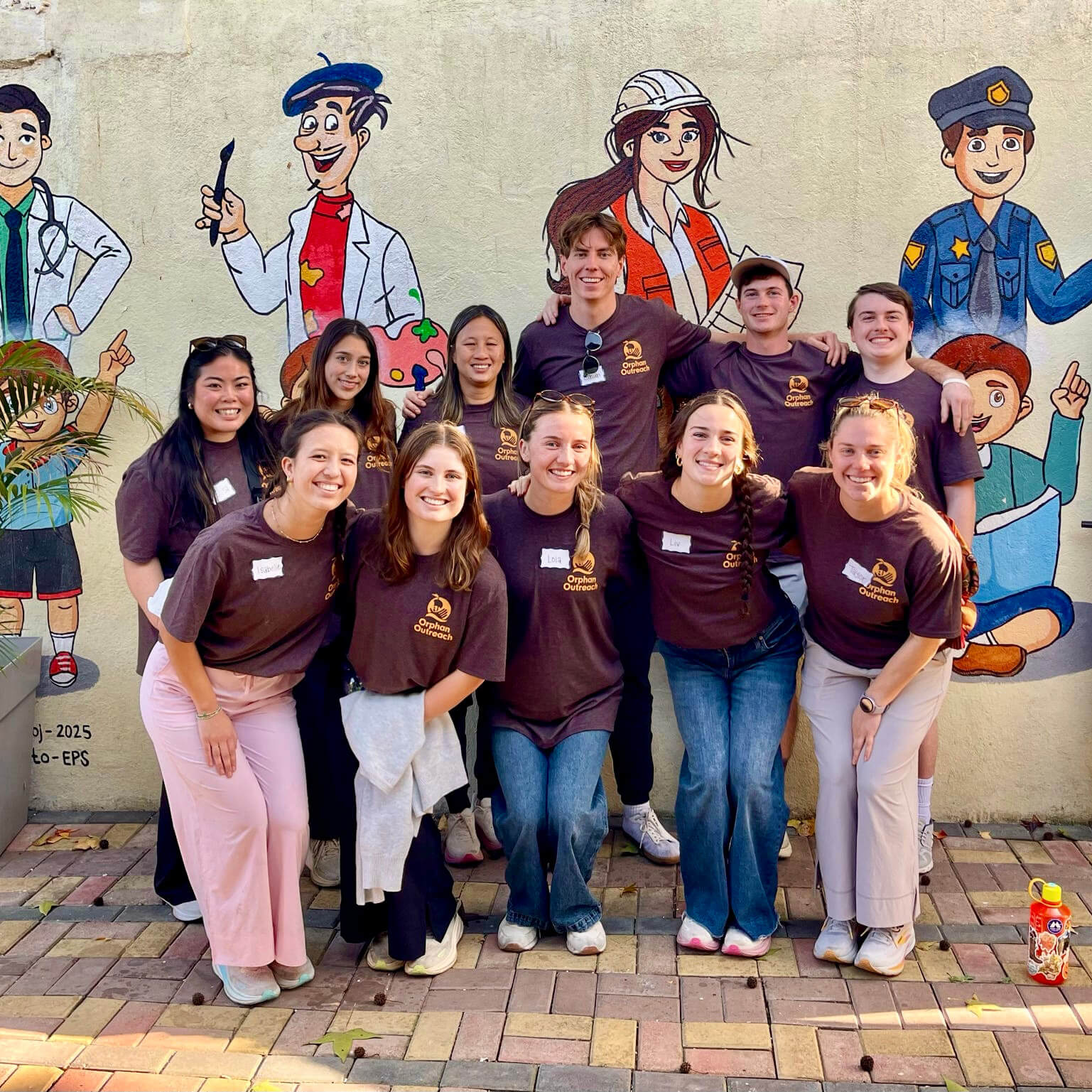 All wearing dark maroon “Orphan Outreach” shirts, Nate Raney poses with other students in front of a concrete wall mural featuring cartoon illustrations of community jobs.