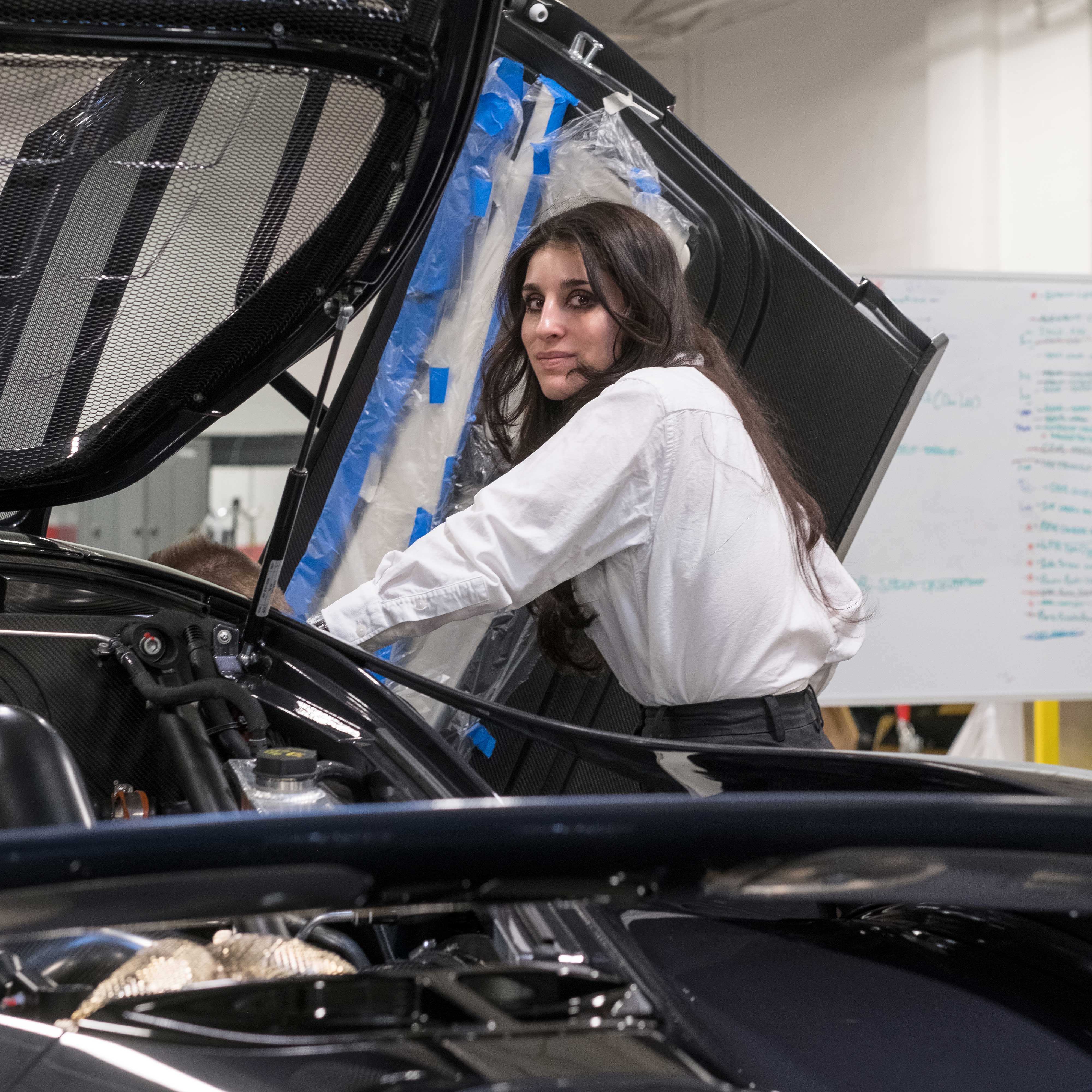 Nazli stands with a car that's being worked on. The hood of the vehicle is open, exposing its engine. She wears a white top.