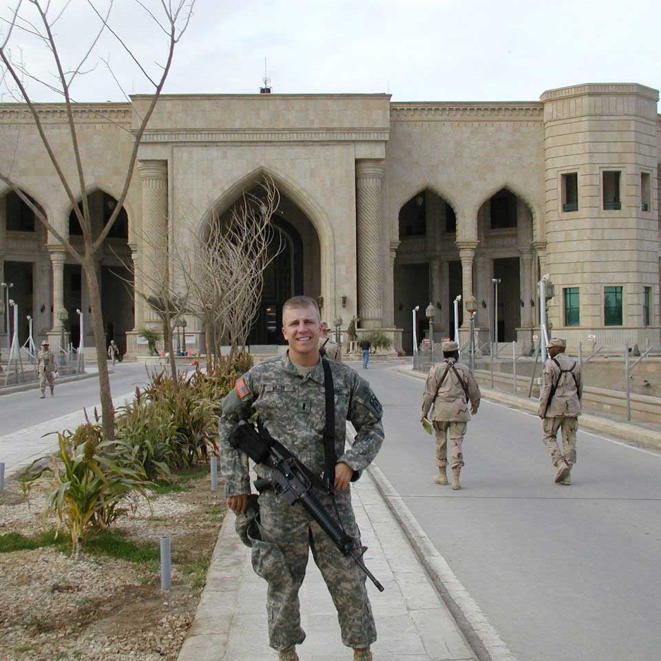 Nick stands outside of a military base. He wears his Army fatigues and has a gun slung across his front. Soldiers mill about.