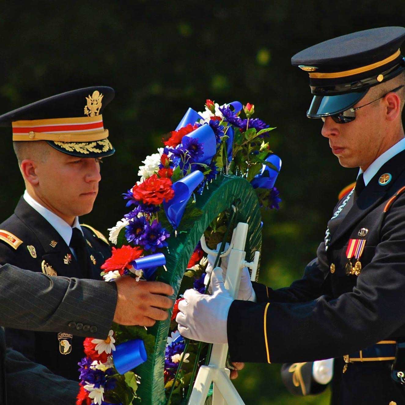 A uniformed Nick lays a hand on a decorated wreath for a fallen service member. Another service member holds the other side. 
