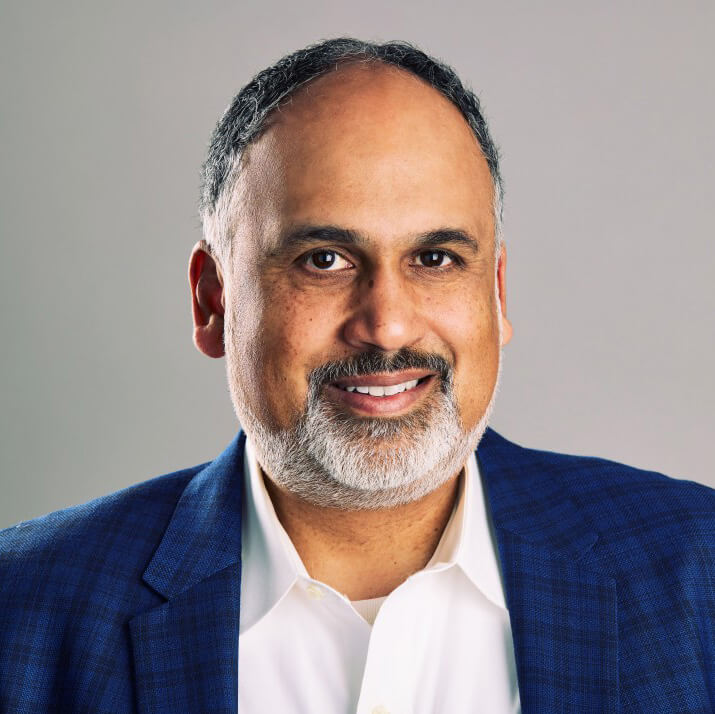 Wearing a blue plaid sport coat, Nishad Pai smiles, posing against a light gray background for a headshot.