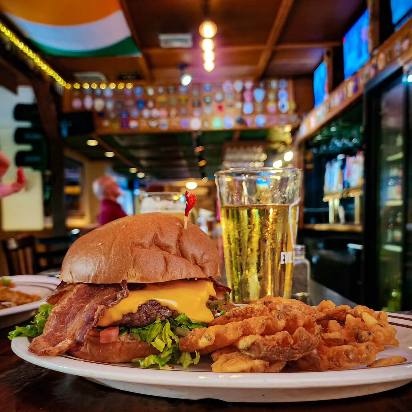 A well dressed bacon burger sits on a plate with fries with a light beer in a glass beside it.