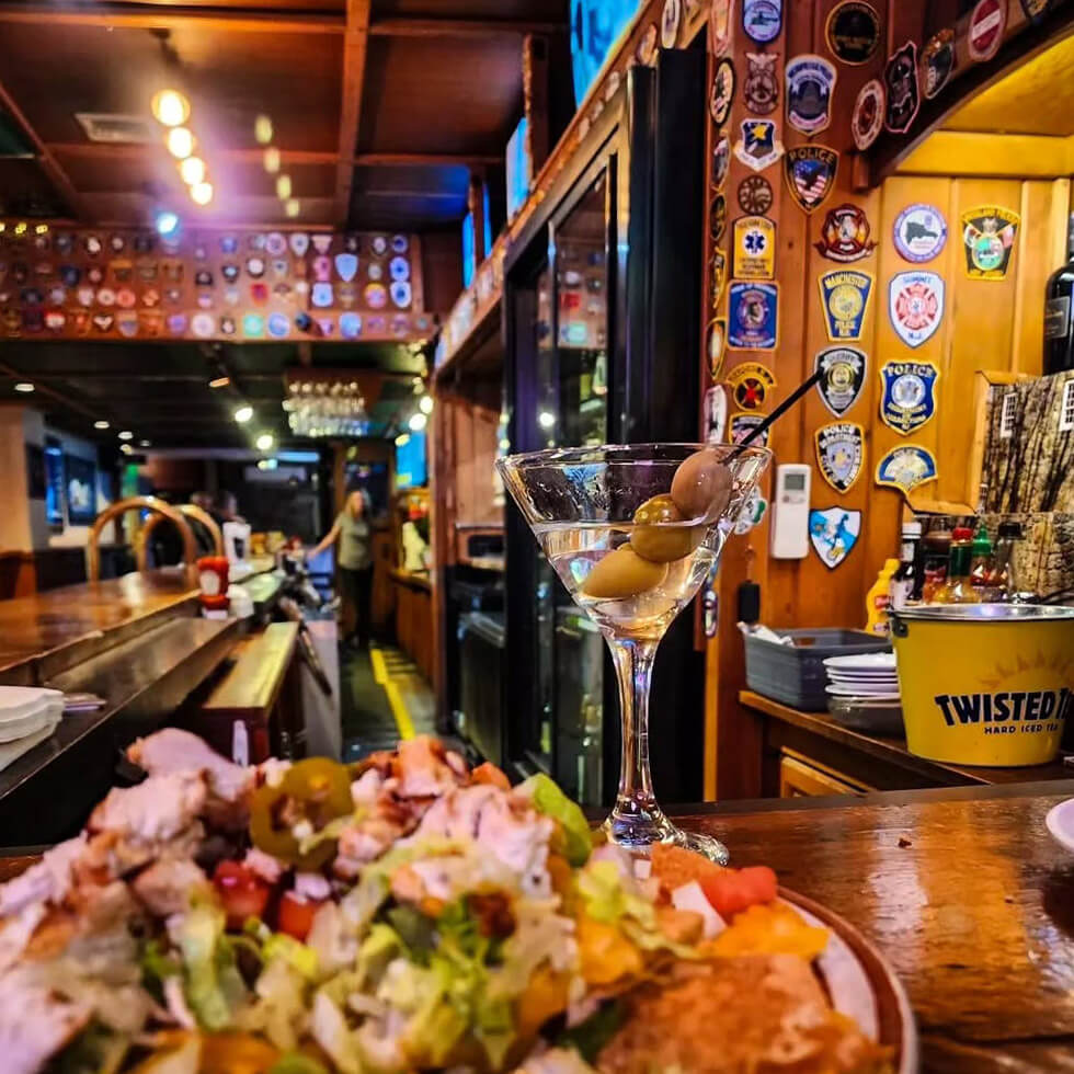 A salad and olive martini are in the foreground on a bartop with hundreds of service patches decorating the walls behind it.