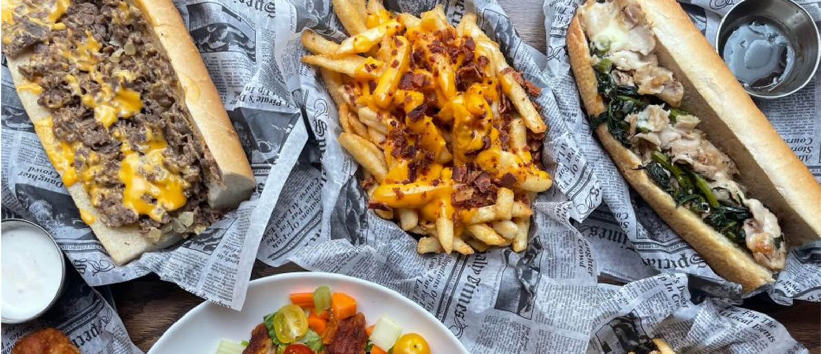 A close up of cheese fries and two cheesesteaks in baskets lined with newspaper print deli paper