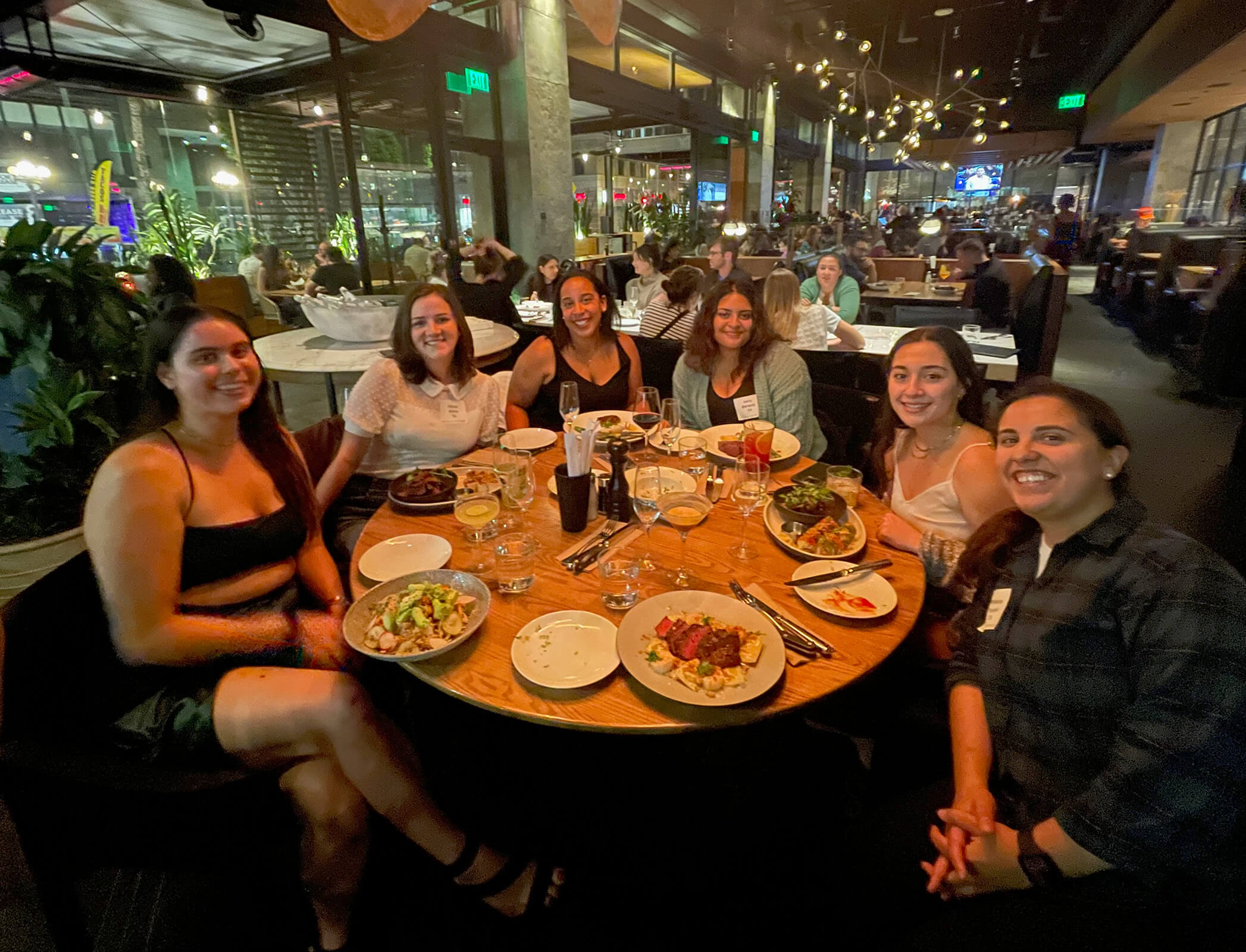 Six women sit at a round table with plates of food in a dimly lit restaurant.