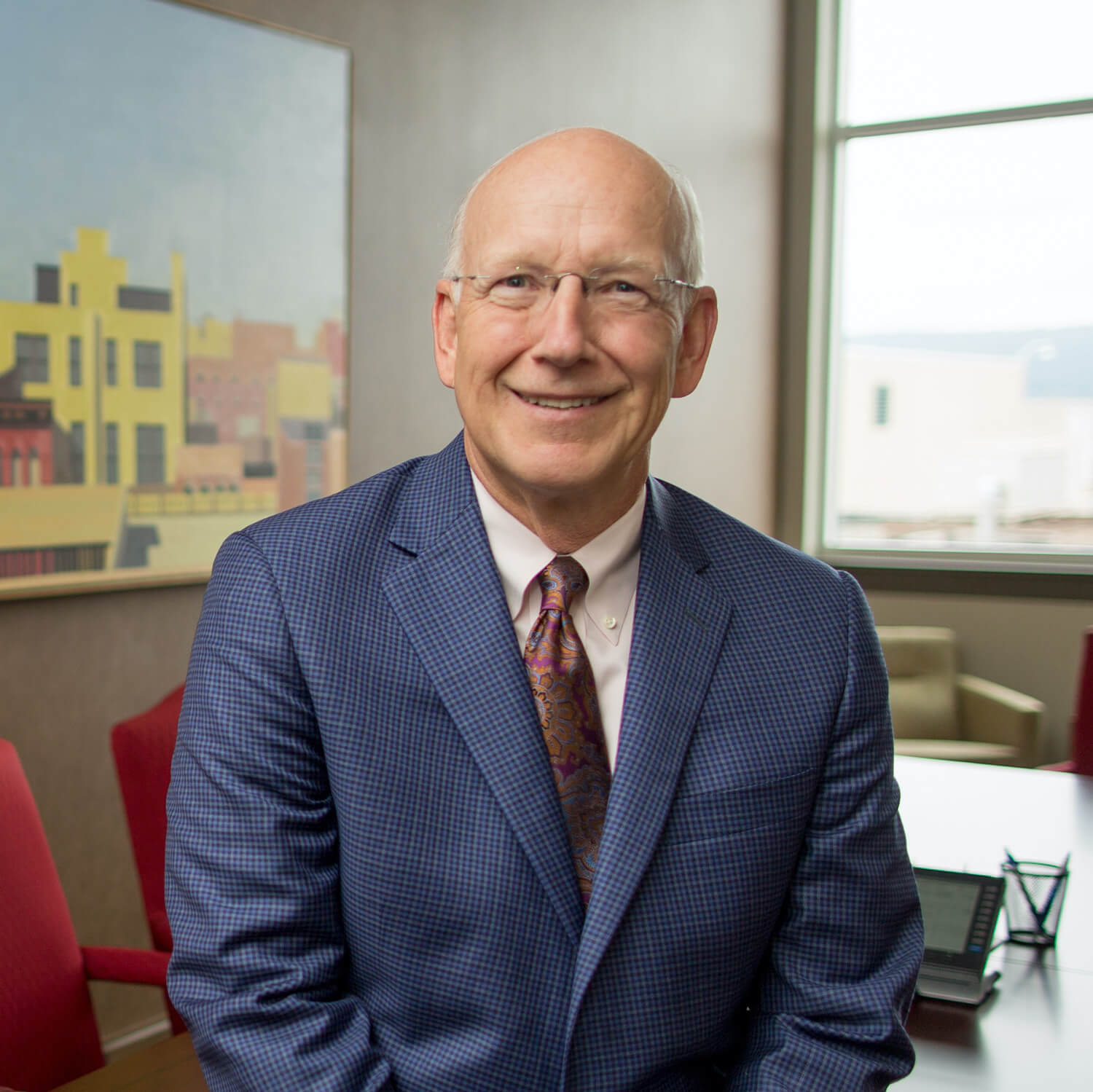 Sitting on the end of a long table in a boardroom, Ollie Foucek wears a blue suit, cream shirt, and multicolored paisley tie and glasses, smiling.