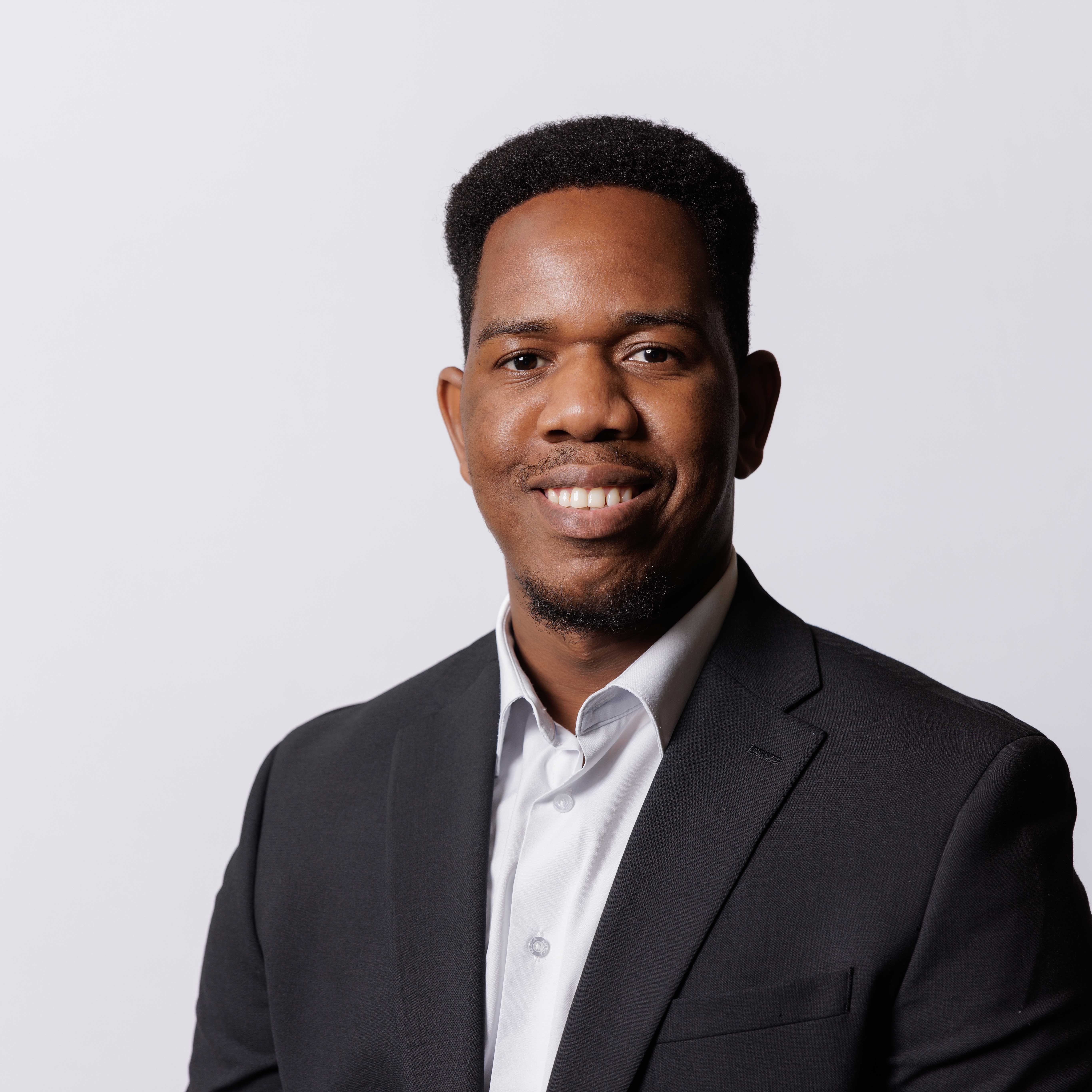 Omogolo wears a black suit jacket with a white button-up underneath and smiles. The room is professionally lit and bright. 