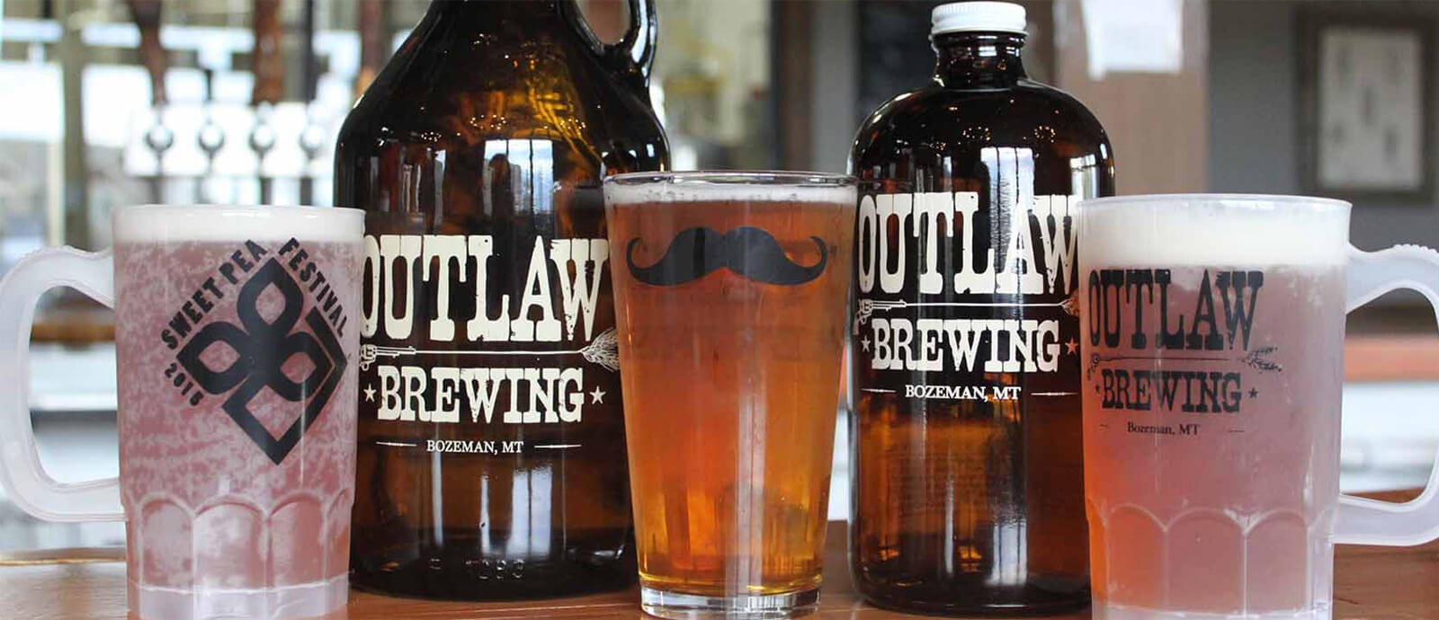 Two dark brown glass jugs with outlaw brewing logos stand between three frosted glasses of beer and foam