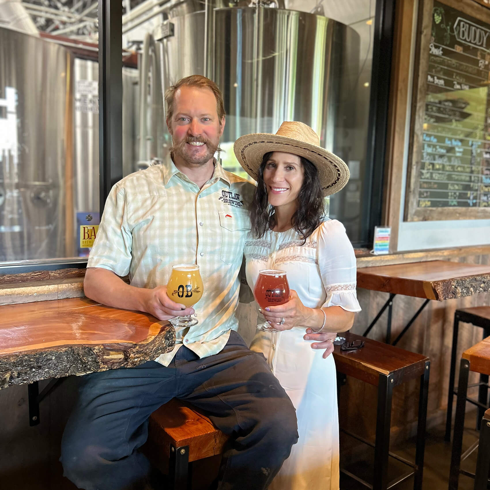 Todd Hough sits on a bar stool at a table made from salvaged wood, while Nicole stands beside him, each holding a glass of beer.