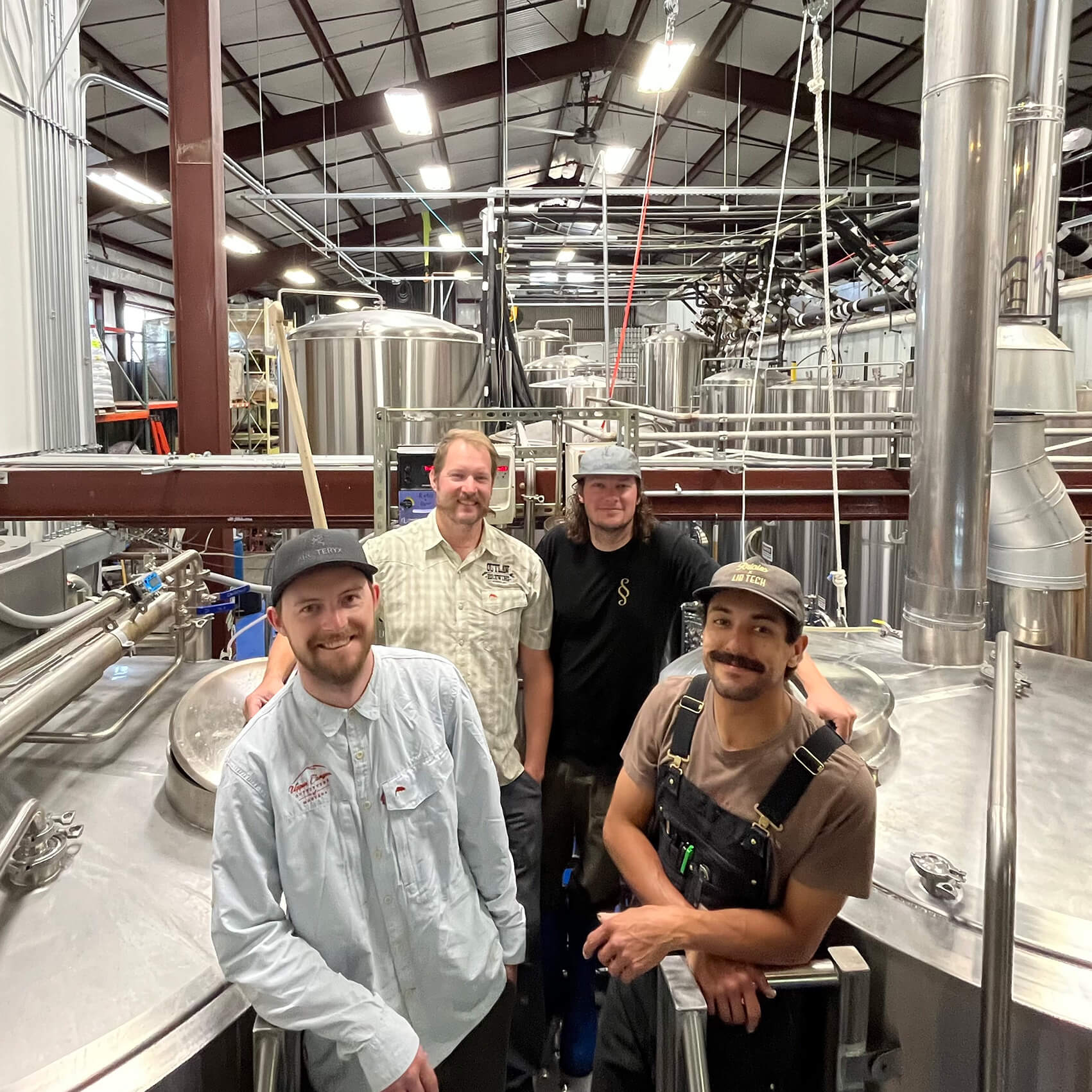 Surrounded by a warehouse filled with metal brewing vats and brown beams, four men gather for a photo.