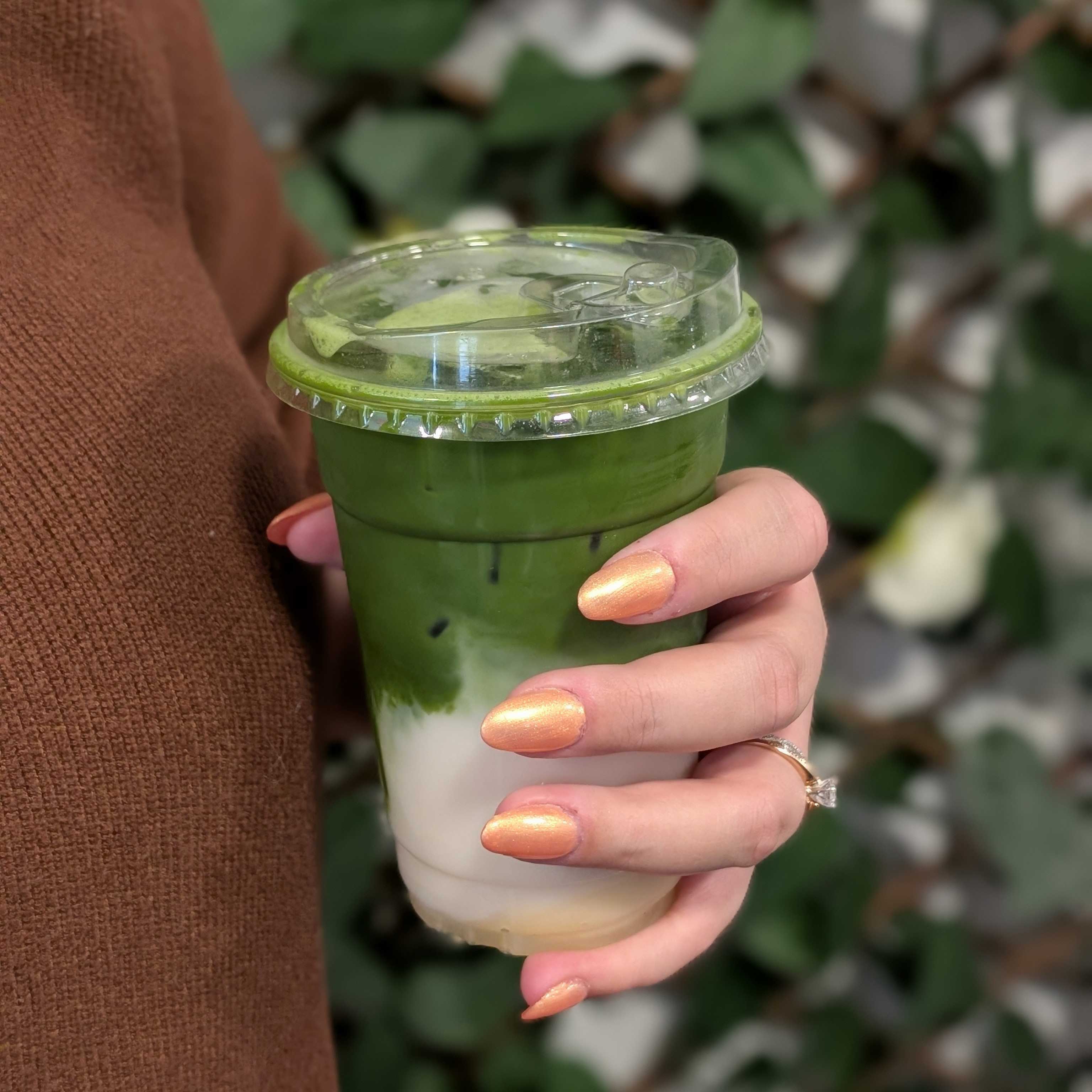 Photo depicts hands holding an iced matcha latte near a wall of greenery. The person's nails are metallic orange. 