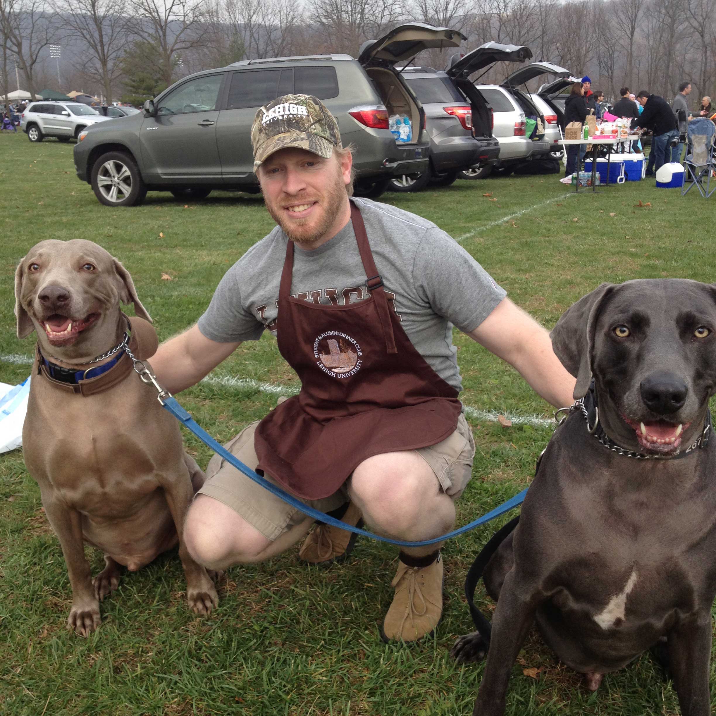Patrick wears a brown Lehigh apron over a grey shirt and a Lehigh hat. He kneels, petting two brown dogs on either side of him.