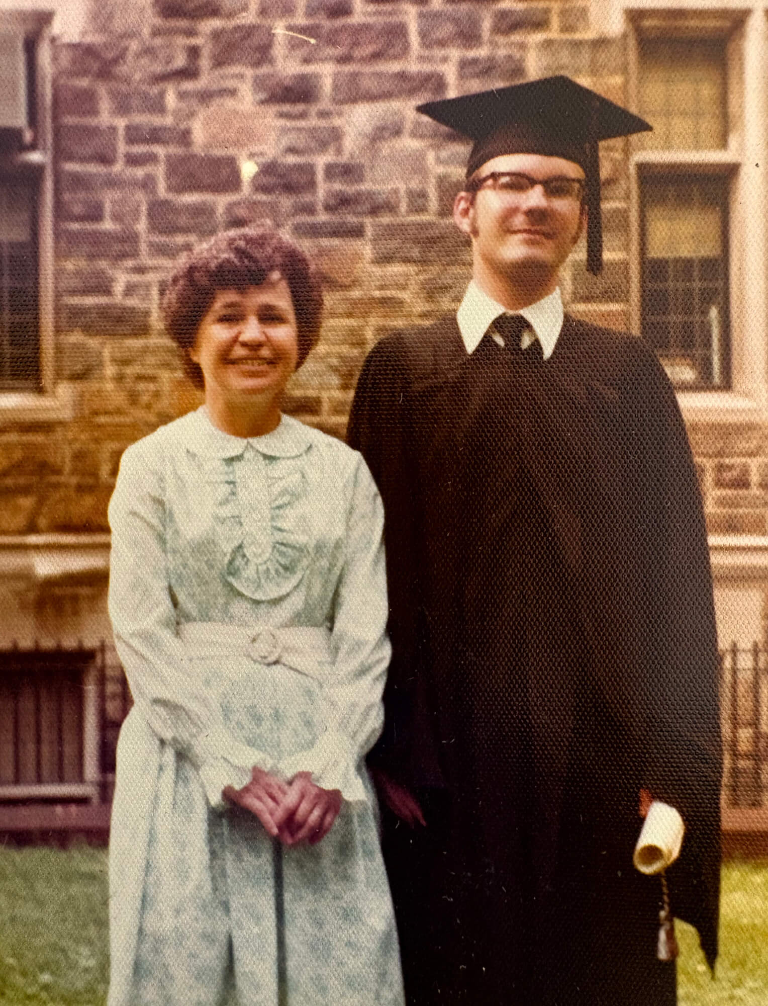 Holding his rolled diploma in his hand, Paul Elosge poses outdoors in graduation cap and gown next to a woman smiling with short dark hair in a powder blue dress.