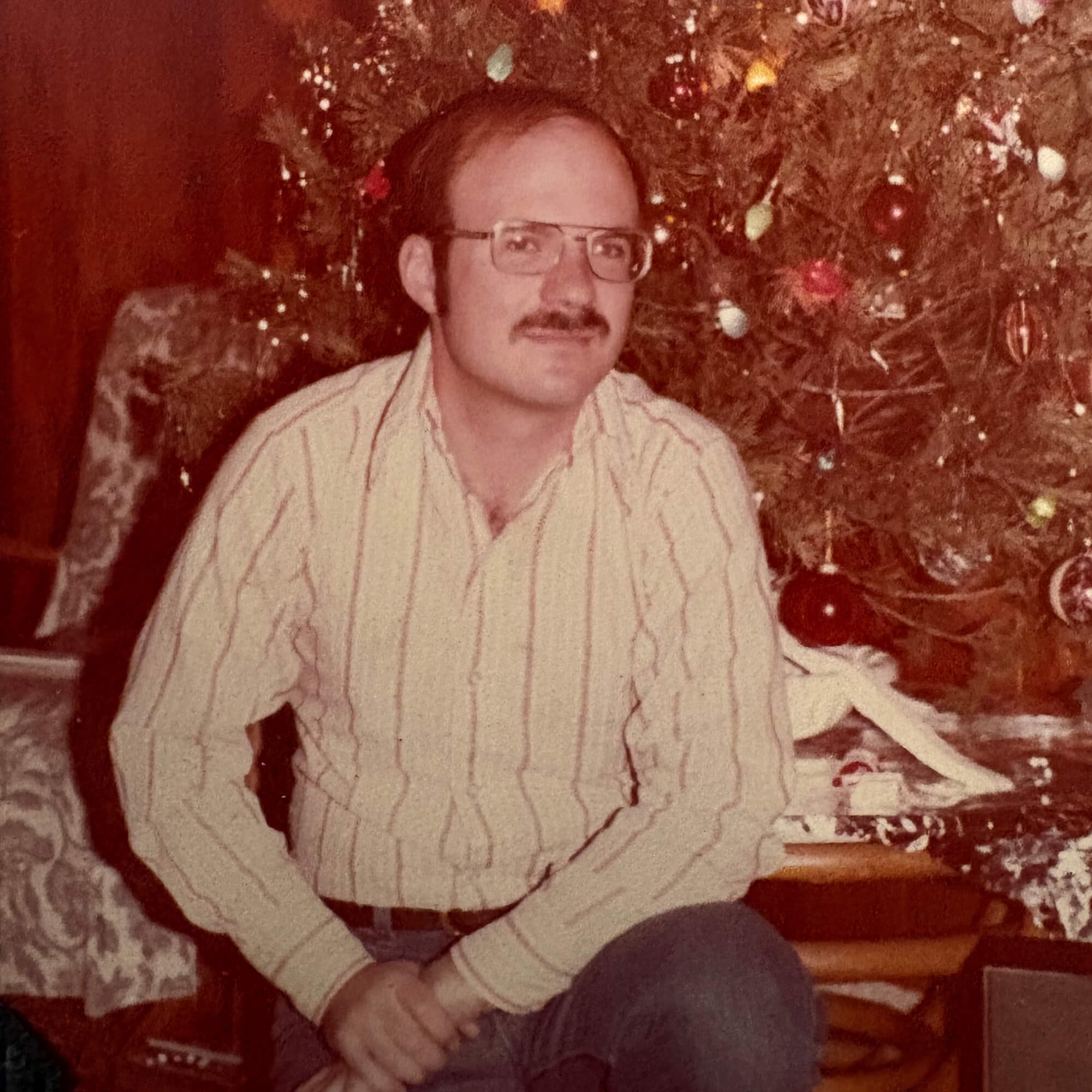 In a photo with colors dulled by age, Paul Elosge leans on one knee in front of a Christmas tree with his hands folded over his knee, wearing glasses and a striped button-down shirt.