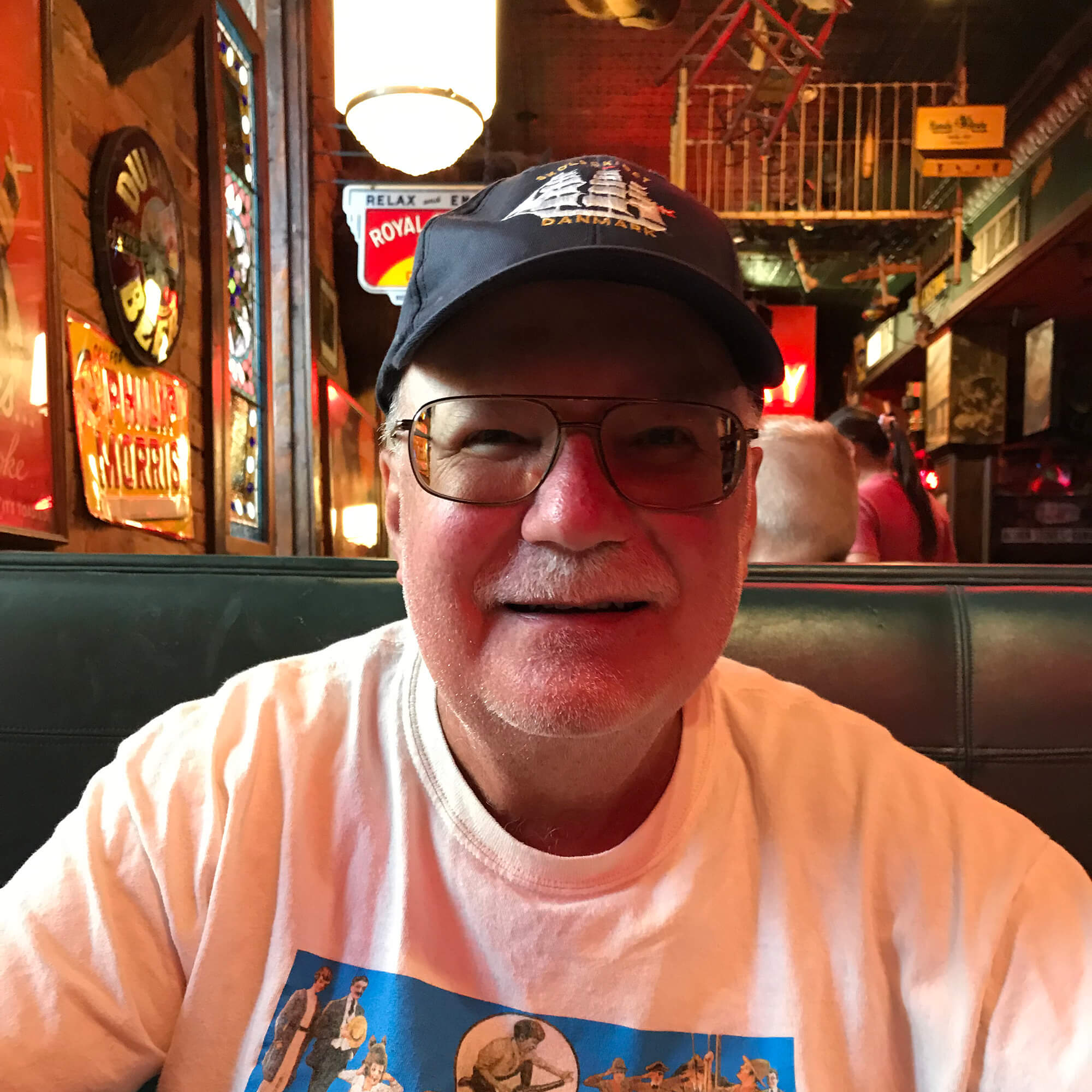 A solo photo of Paul Elosge sitting in a restaurant booth, wearing a baseball cap and t-shirt, with pub-style decor in the background.