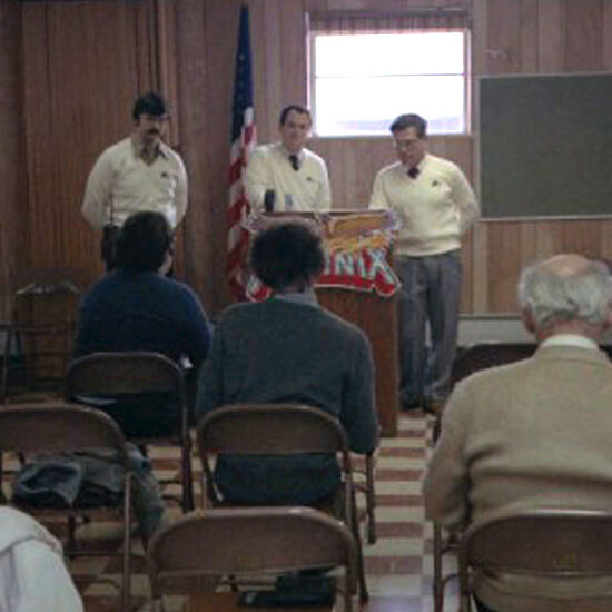 Three men stand in a wood panelled room speaking at a podium to some men assembled in folding chairs.