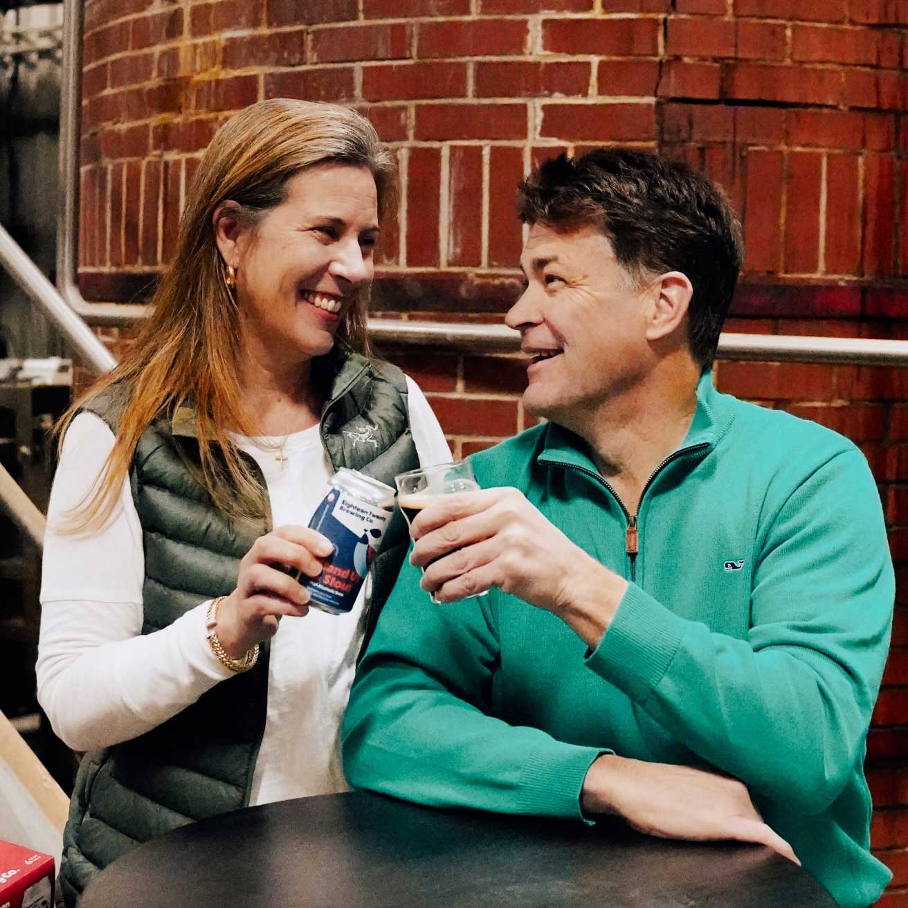 Robin and Alan clink their glasses together; Robin holds a can of 1820 Brewing Co. while Alan holds a pint of beer.