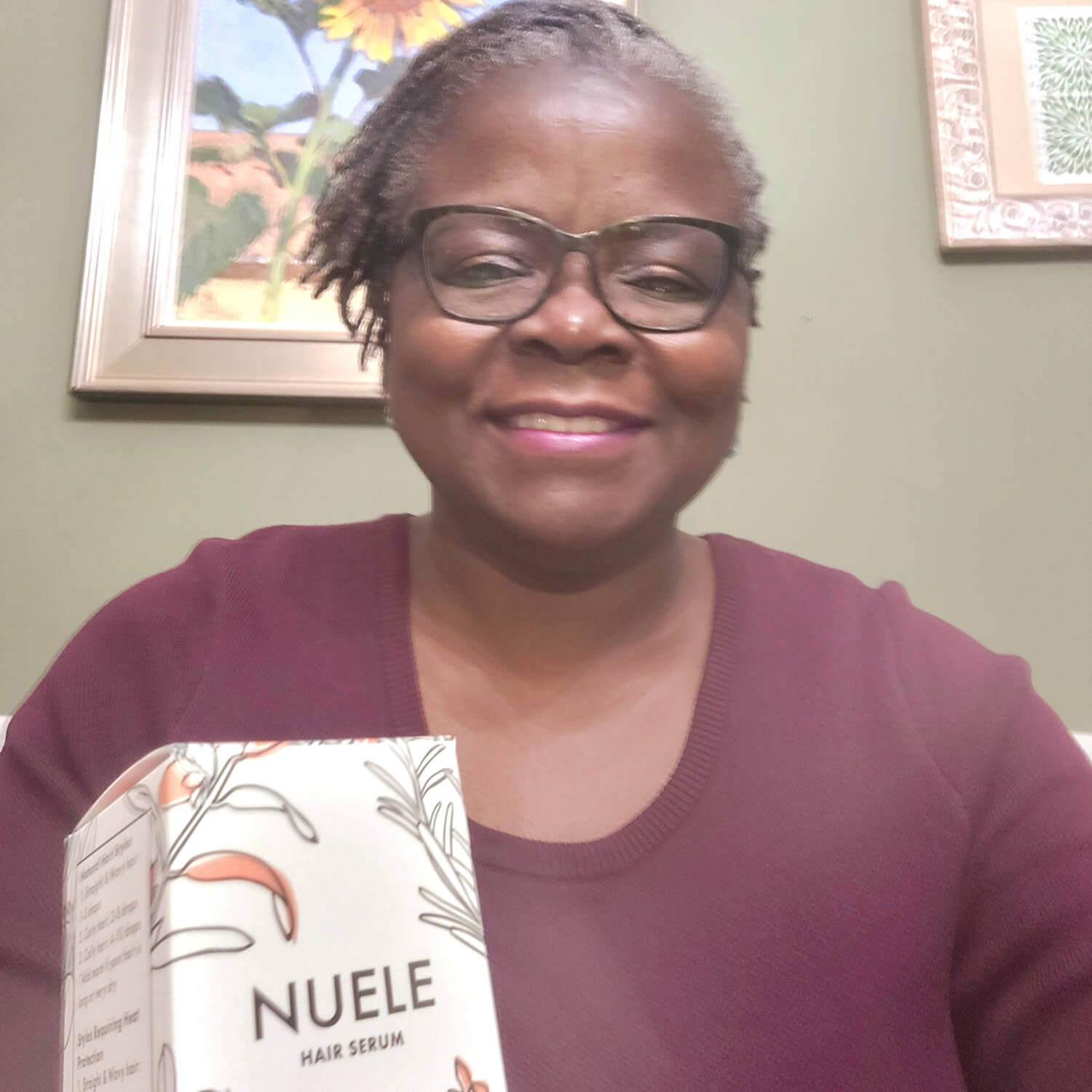 Martey-Ochola smiles while wearing a maroon sweater and black glasses. In front of her is the Nuele Hair Serum.