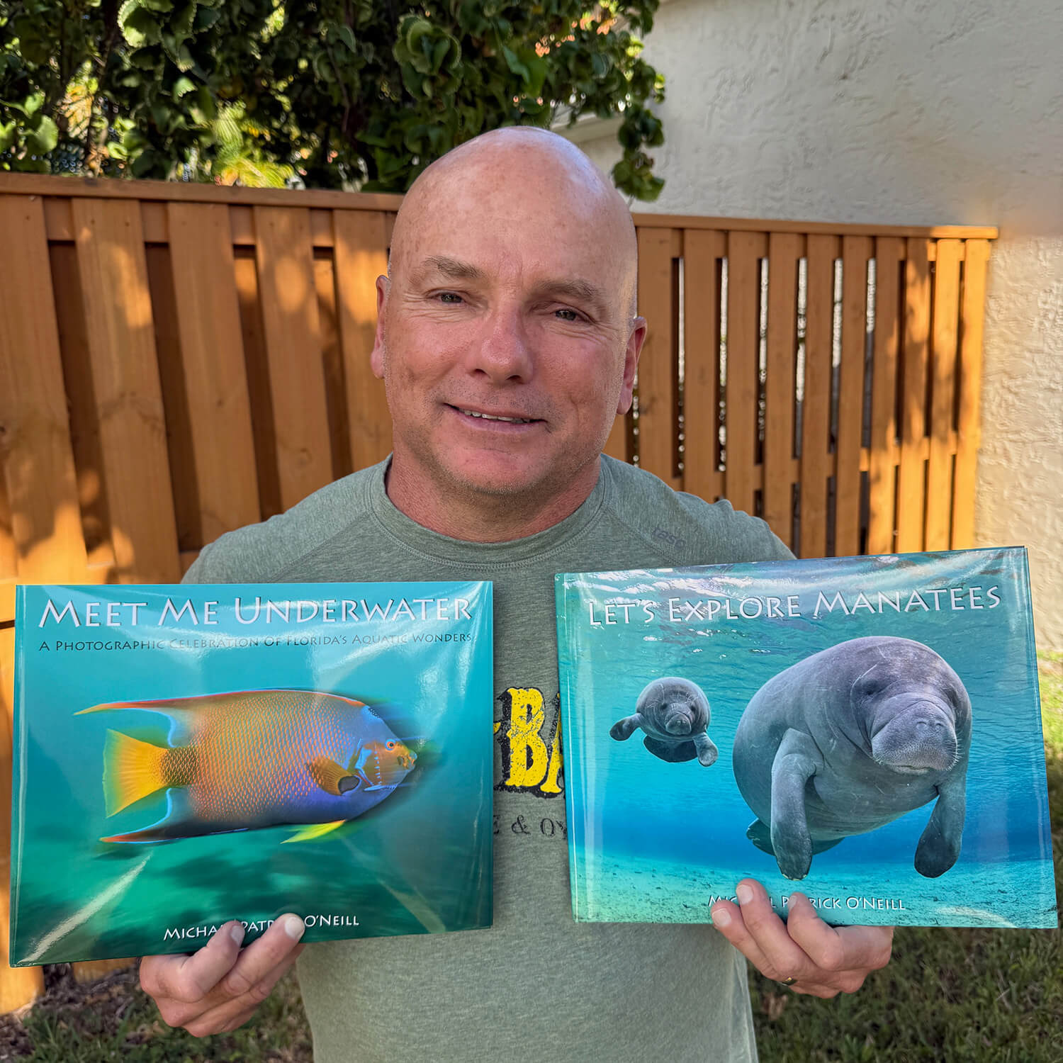 Michael stands and holds two of his books. In his left hand: ‘Meet Me Underwater’ and in his right: ‘Let’s Explore Manatees’.