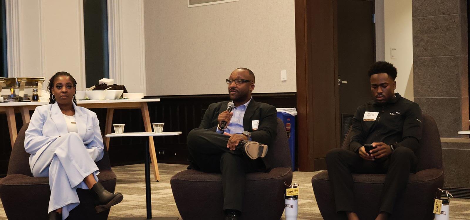 Noel, wearing a black suit jacket and slacks, holds a microphone and chats on a panel between two other Lehigh alums. 