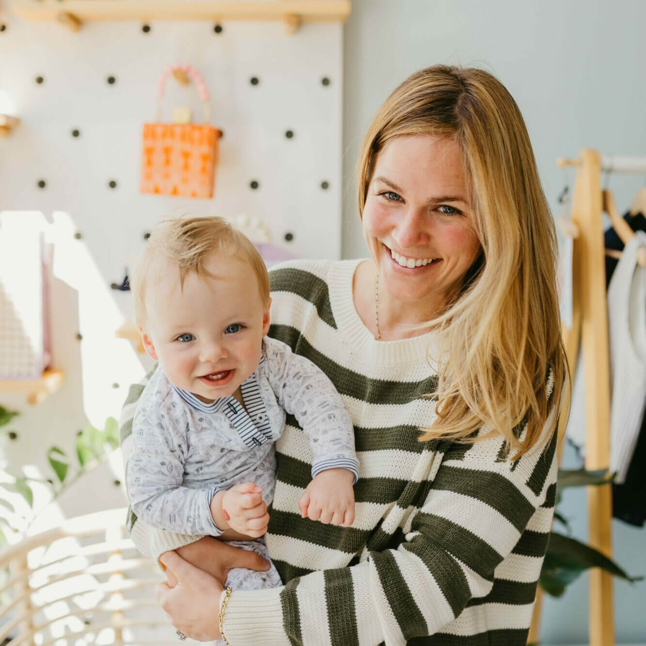 Ferrel wears a striped sweater and holds a smiling baby that’s wearing a set of her pajamas in a bright, modern room. 