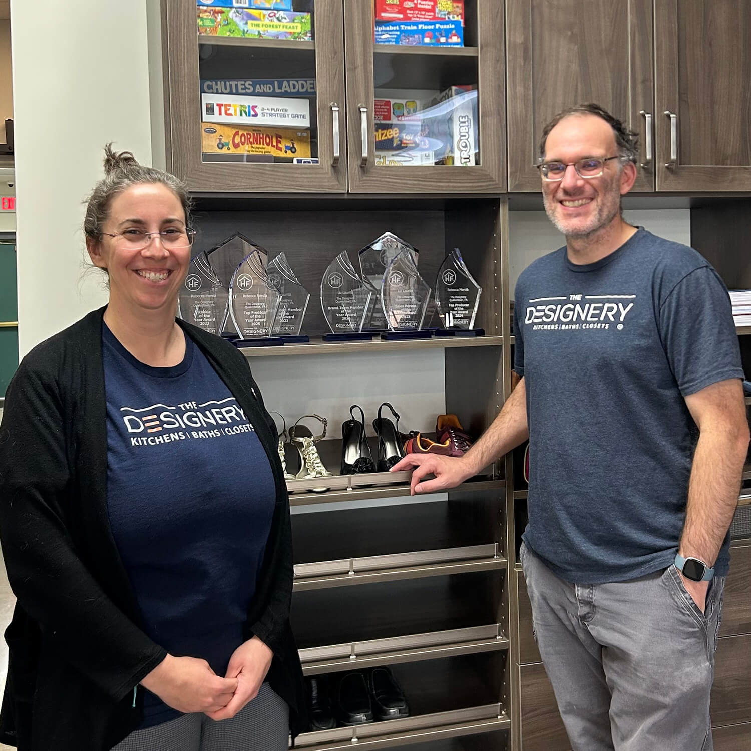 Rebecca and her husband wear shirts with the business name; behind them are dark cabinets and several awards the company has won.