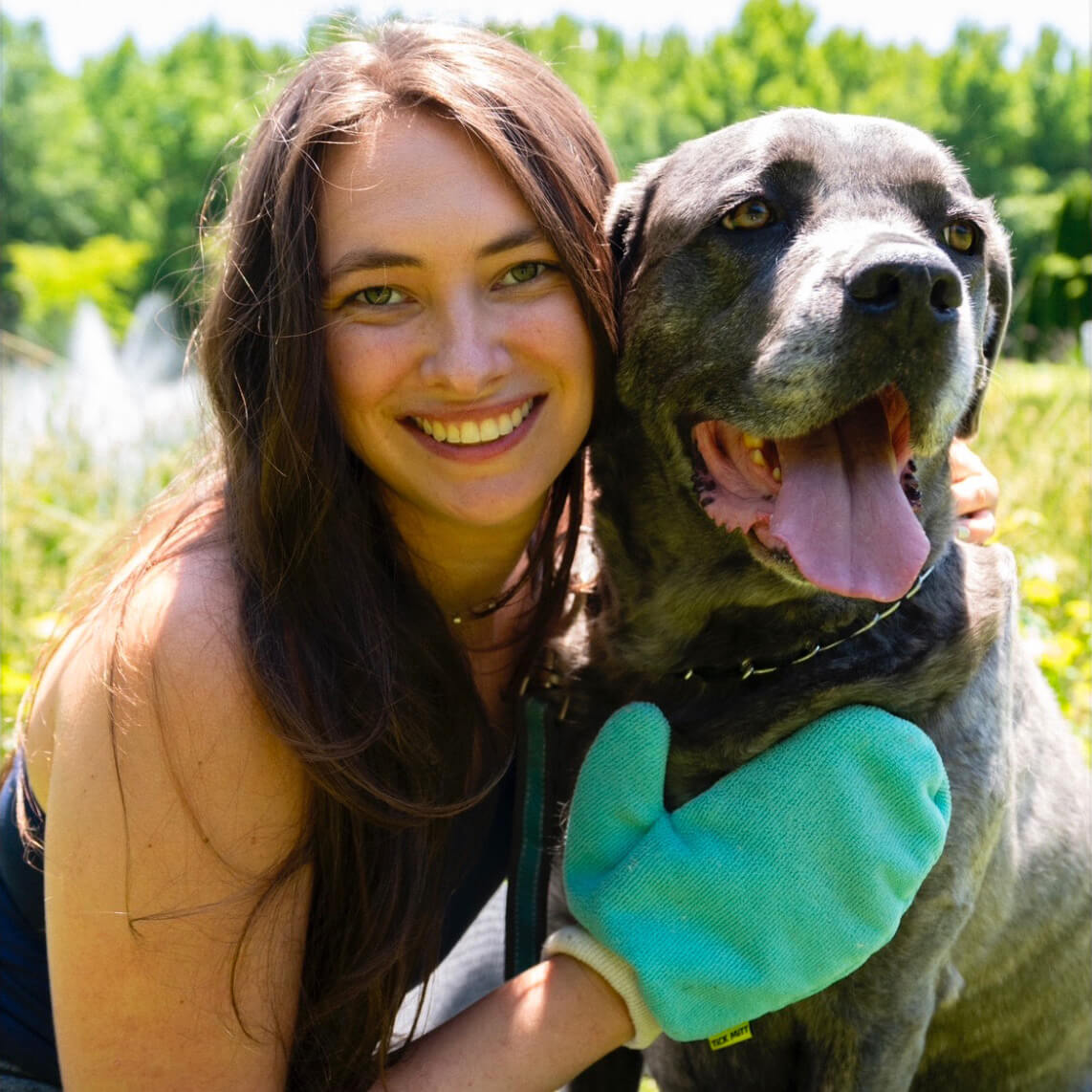 Olivia sits in the grass alongside a large grey dog. She has a blue TiCK MiTT on her hand and rests that on the dog’s chest.