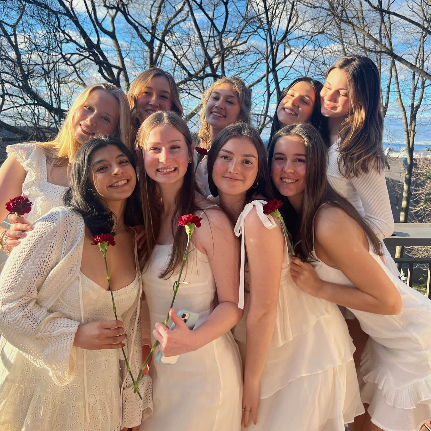 Nine sisters from Pi Beta Phi sorority pose, each wearing a white dress and holding a single red carnation.