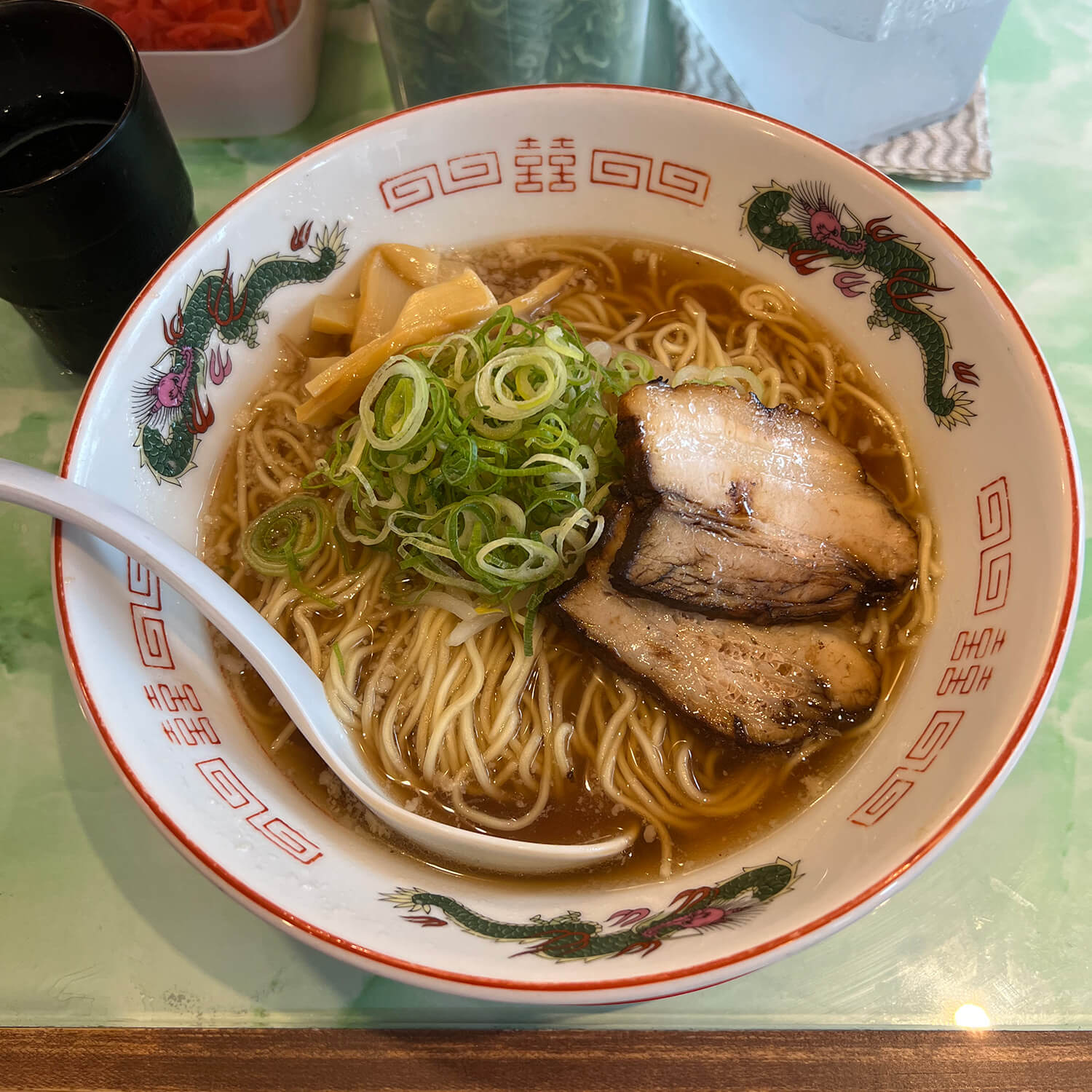  A white porcelain bowl with dragons painted on the rim filled with ramen, meat, and green onions.