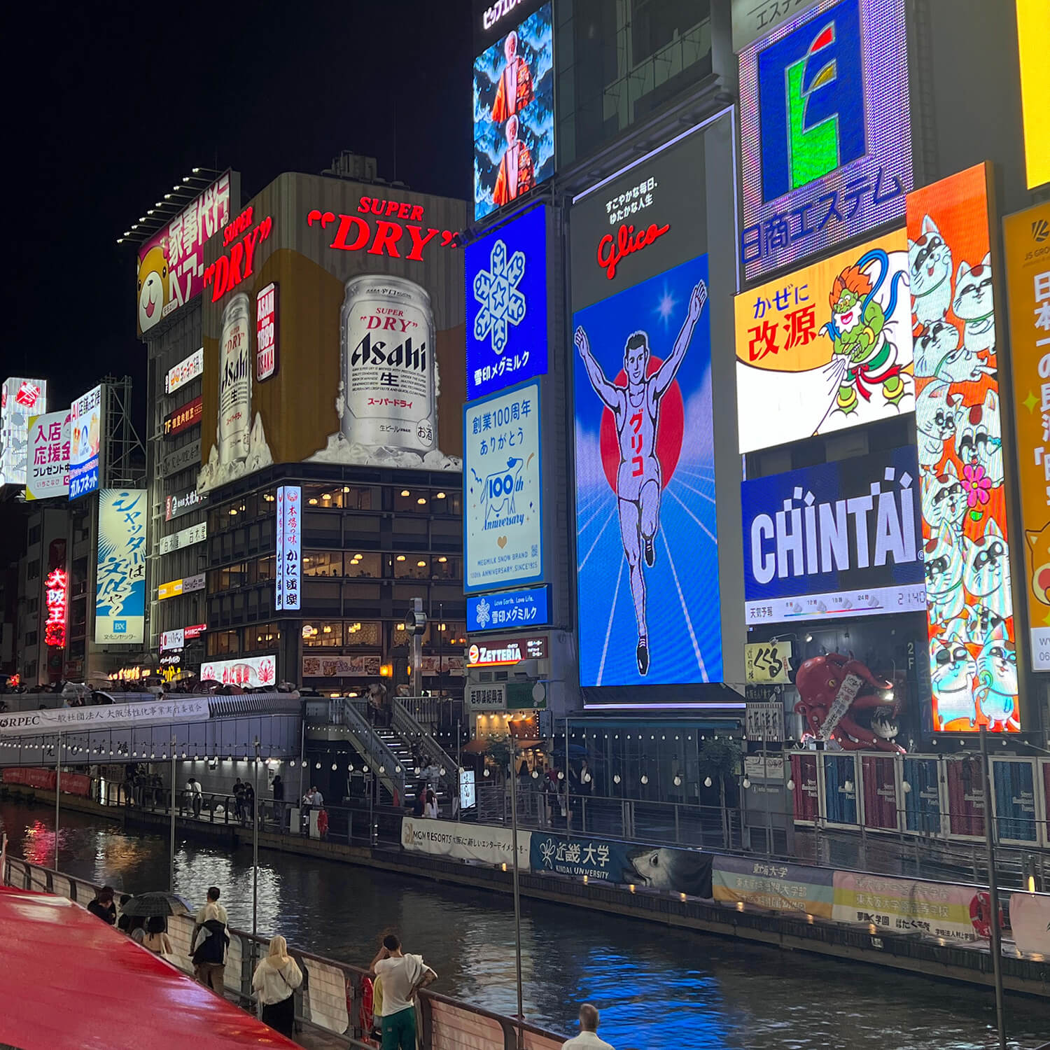 A riverbank lined with tall digital billboards at night in Japan, with the center one featuring an illustration of a track athlete, arms raised in victory under a “Glico” logo.