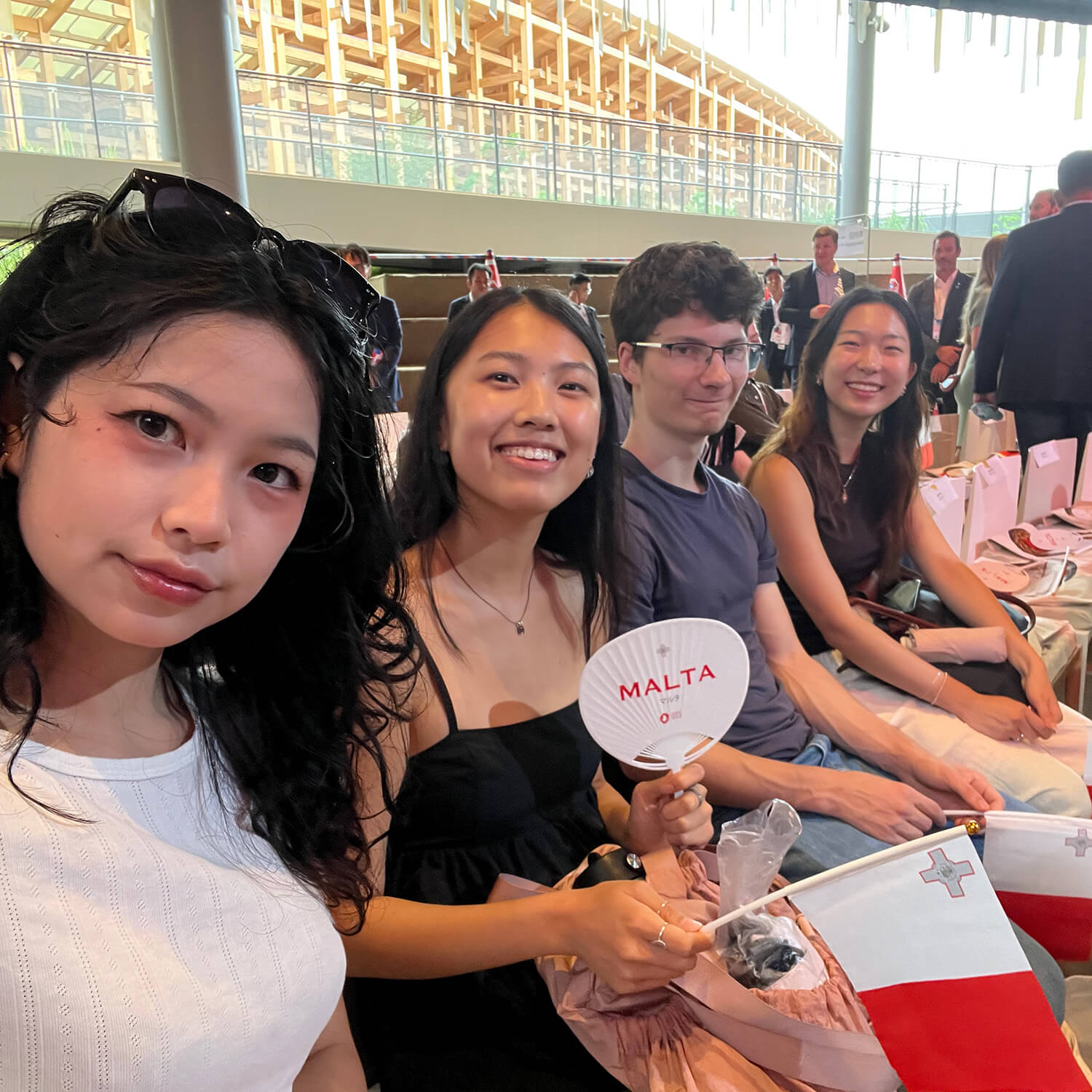 Four students sit on chairs positioned beside each other as one closest to the camera takes a selfie and others hold red and white flags.