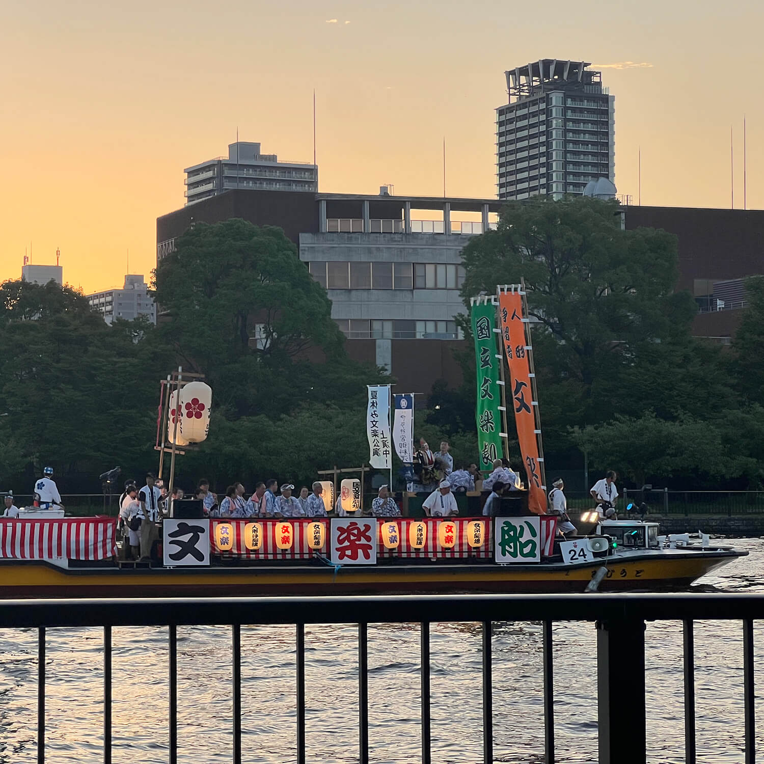 At sunset along a river, a small barge decorated with Japanese flags and lanterns floats with buildings in the distance.