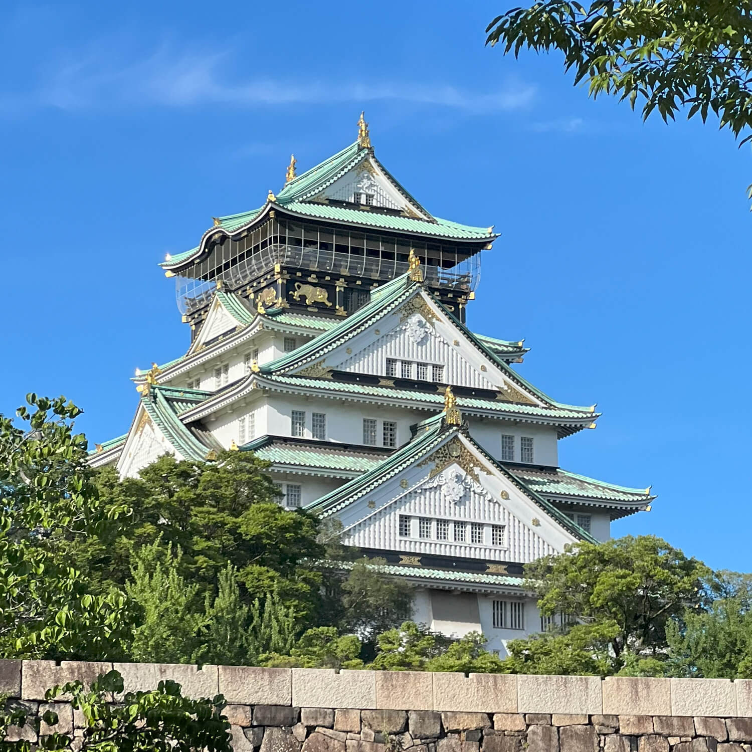 A Japanese style castle with gold embellishments, white siding, and mint green roof surrounded by green trees.