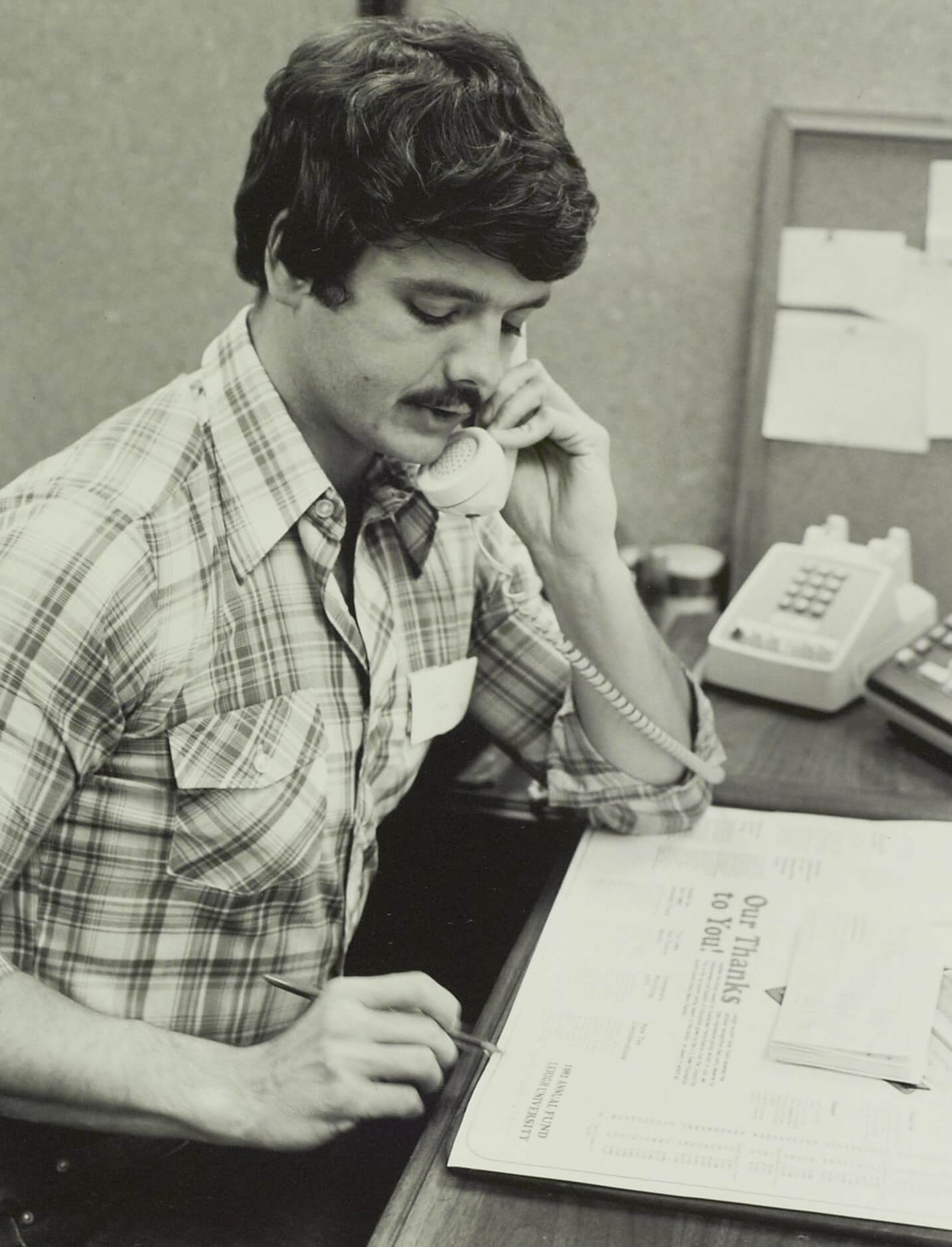 In a plaid button-down shirt with sleeves rolled up, Paul Elosge leans over a desk on the telephone holding a pen.