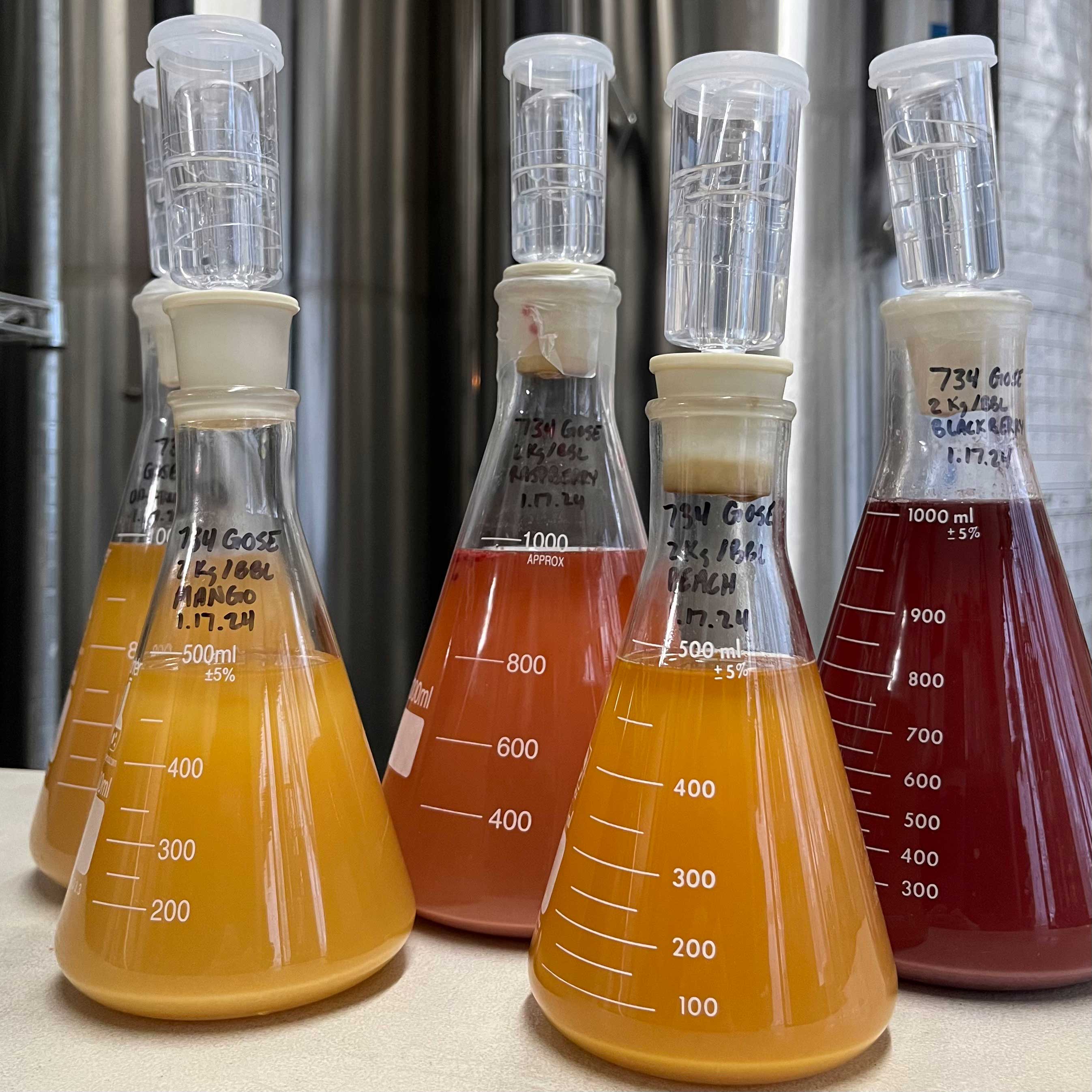 Four triangle beakers hold liquid in varying shades of red and orange. Technical specs and the names of fruit are written on them.