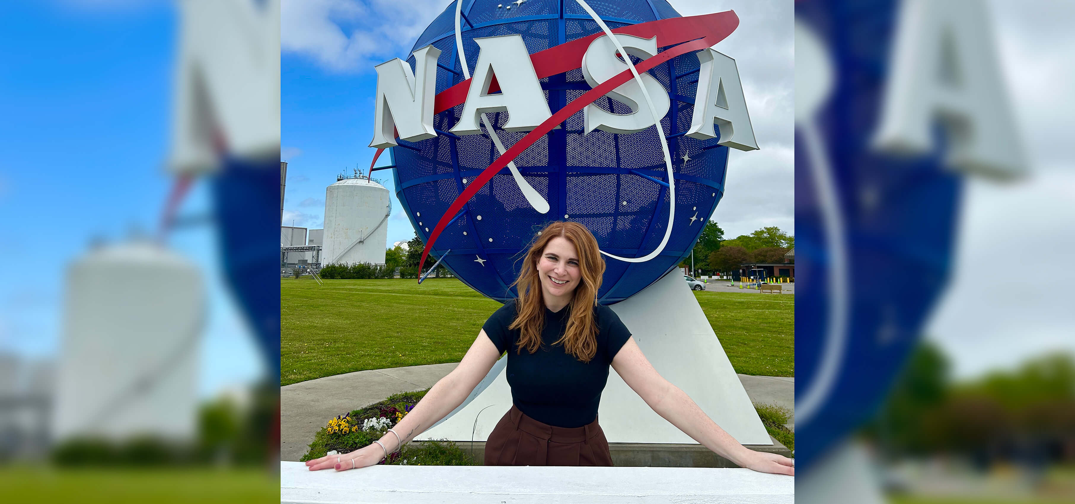 A woman stands smiling in front of a large NASA logo sculpture outdoors on a grassy area with blue sky and clouds.