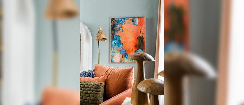 The corner of a living room with a colorful abstract painting, bright furniture, pillows, a lamp, and wooden mushroom figurines.