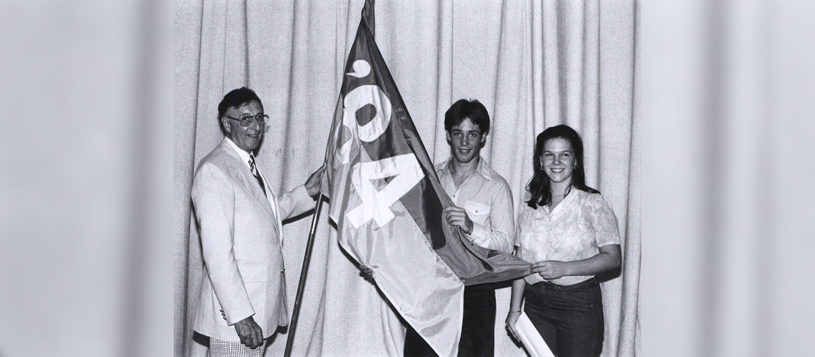 A black and white photo of a man in a light colored suit standing with two students on stage, all holding a large flag together that reads “’84.”