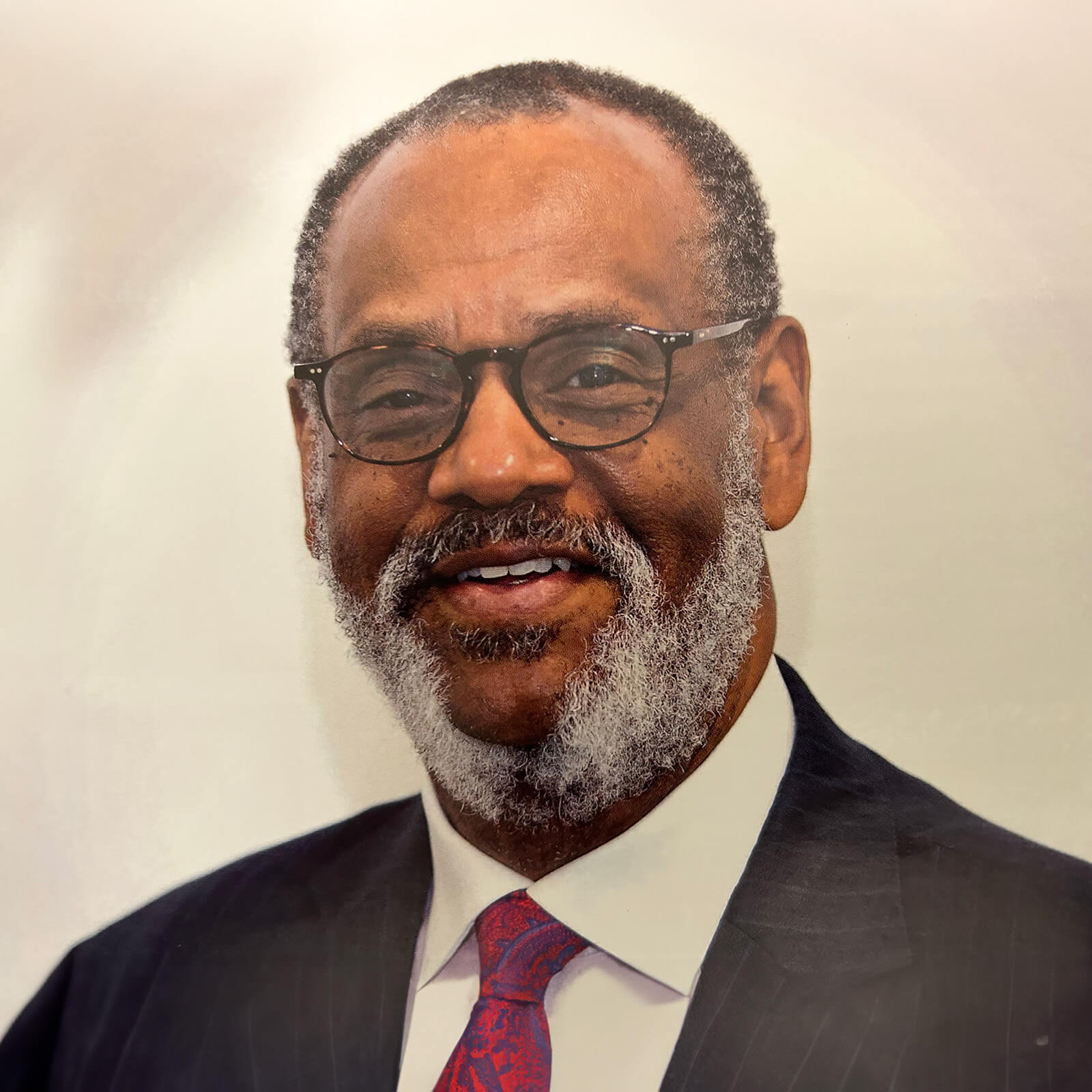 Ralph Thomas poses for a headshot while smiling at the camera and wearing glasses, suit, and red patterned tie.