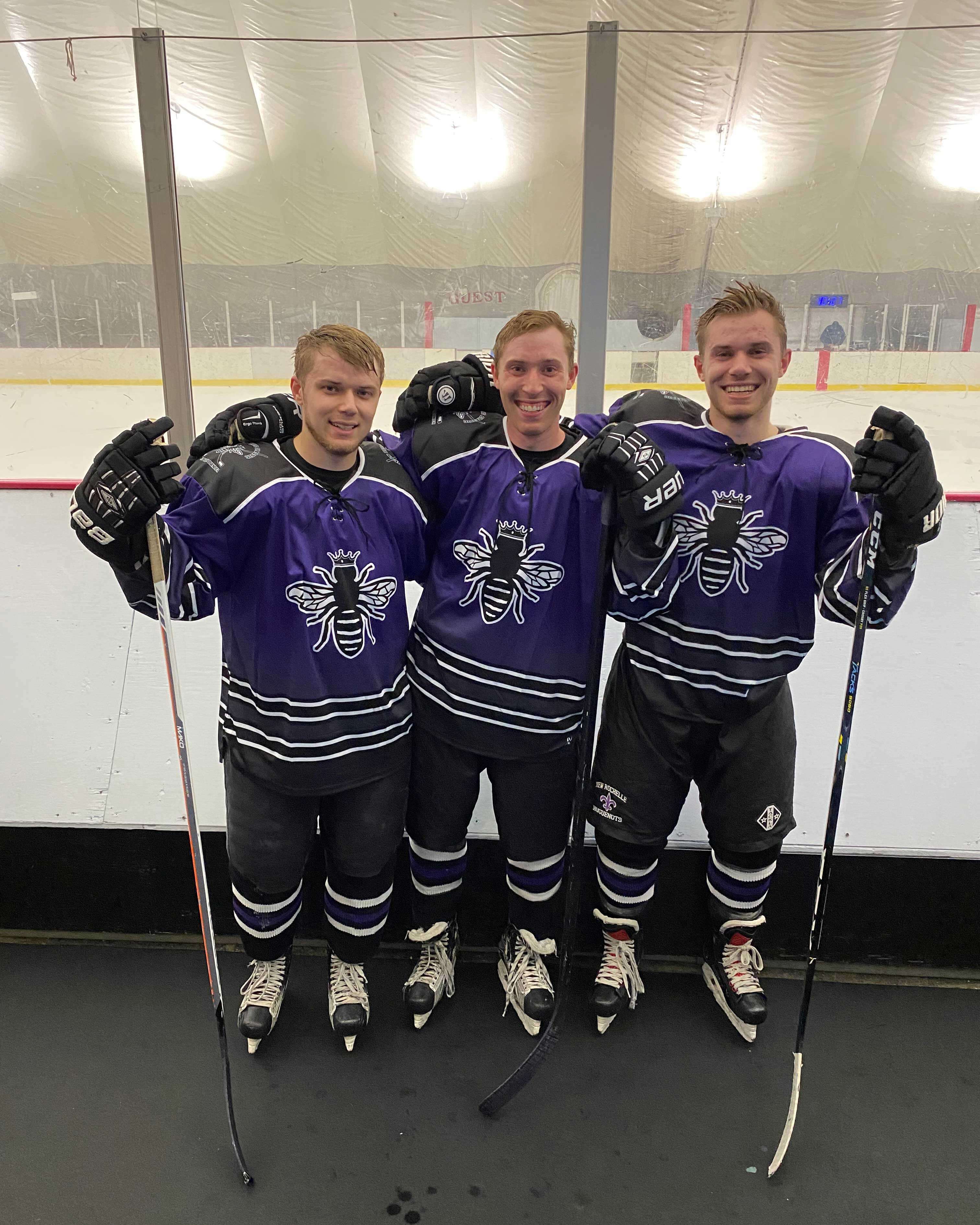The brothers stand together after an ice hockey game. They each wear their purple jerseys and hold a hockey stick in gloved hands.